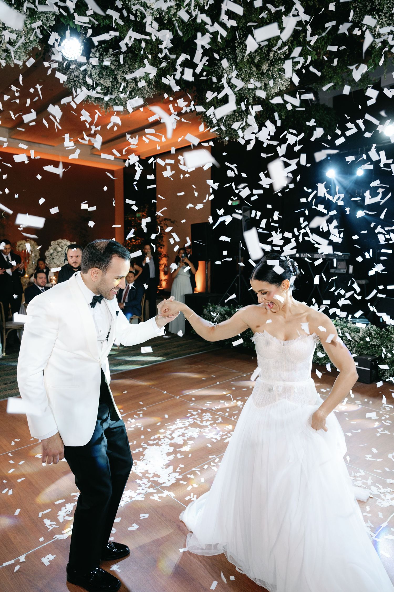 Newlyweds share a joyful dance surrounded by falling white confetti in a beautiful reception hall.