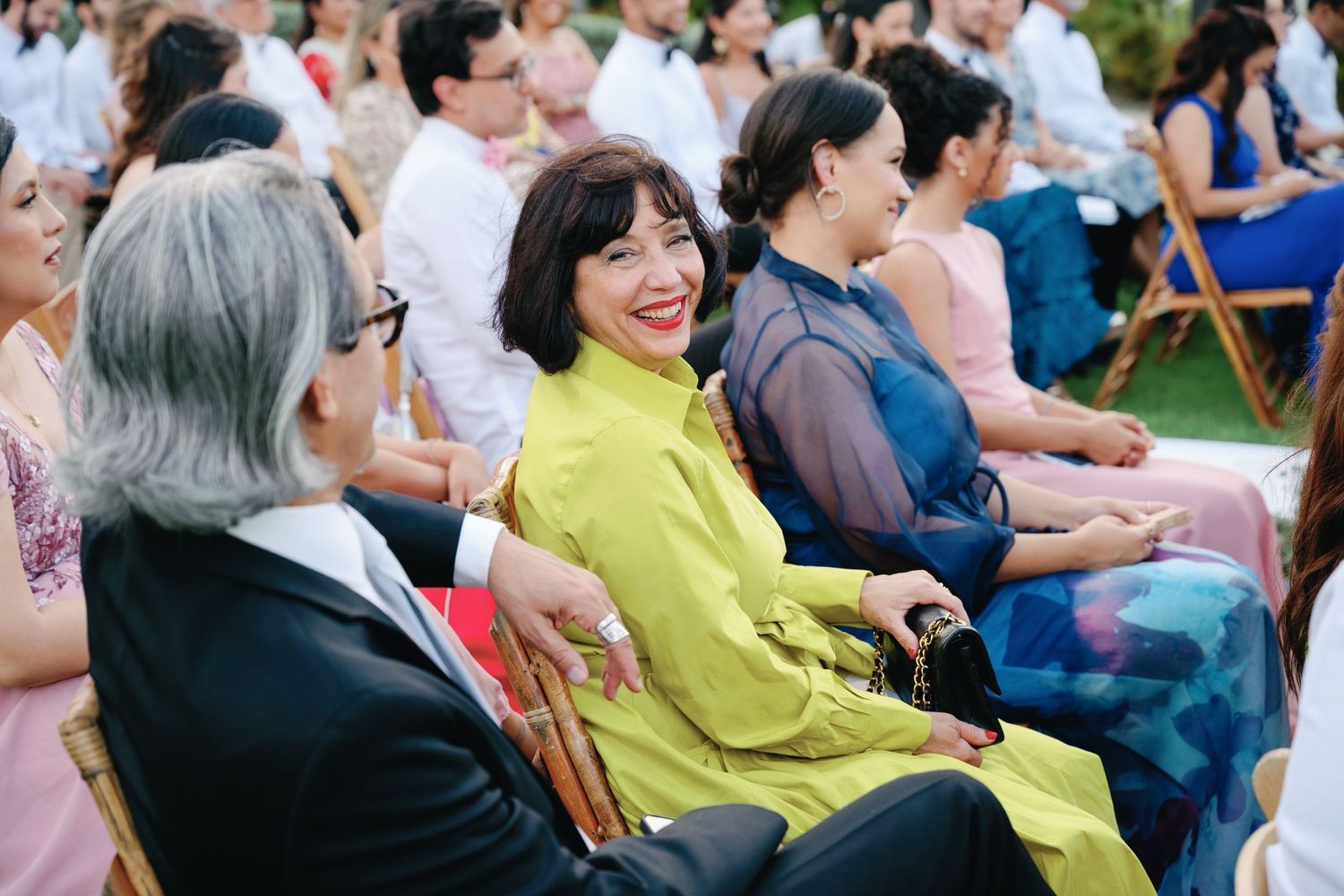 Wedding attendees watching ceremony from wooden chairs arranged outdoors under palm trees.