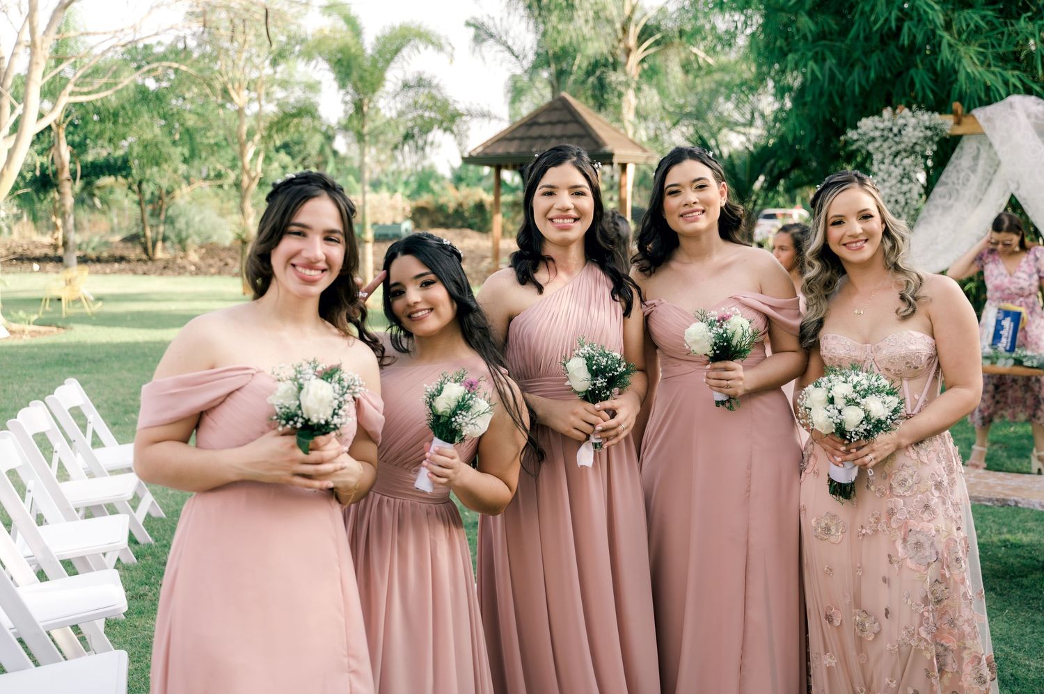 Bridesmaids in dusty rose pink dresses hold white bouquets while standing together at an outdoor garden wedding ceremony.