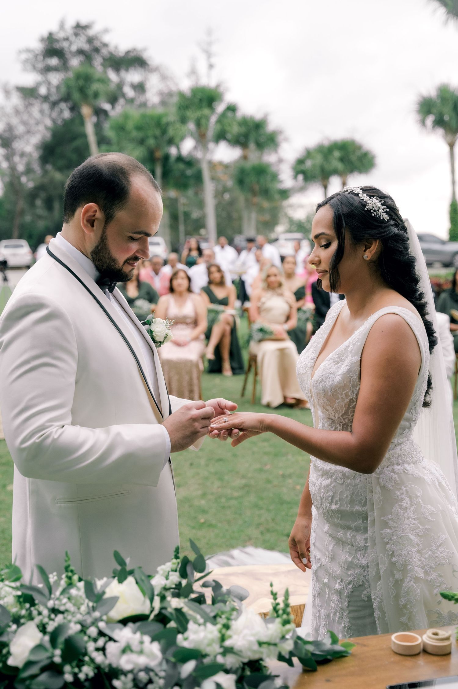 A couple shares a tender moment during their wedding ceremony, exchanging rings outdoors on a sunny day.