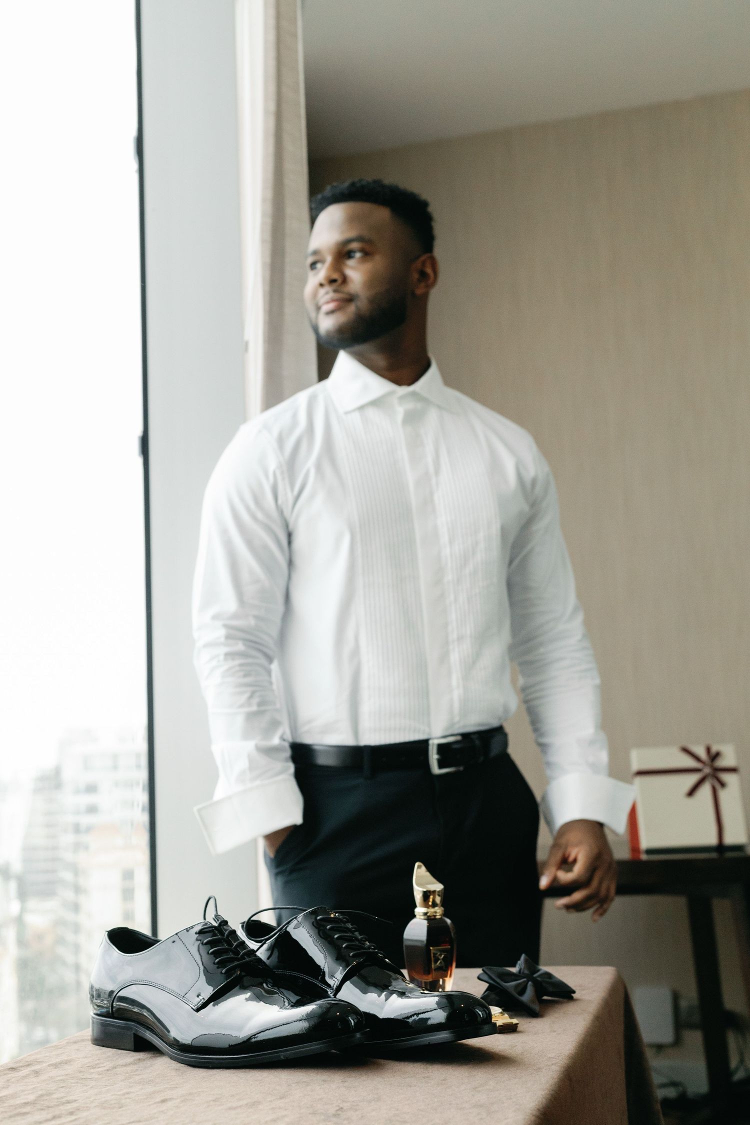 A person in a crisp white dress shirt and black pants stands near a window with formal shoes displayed on a table.