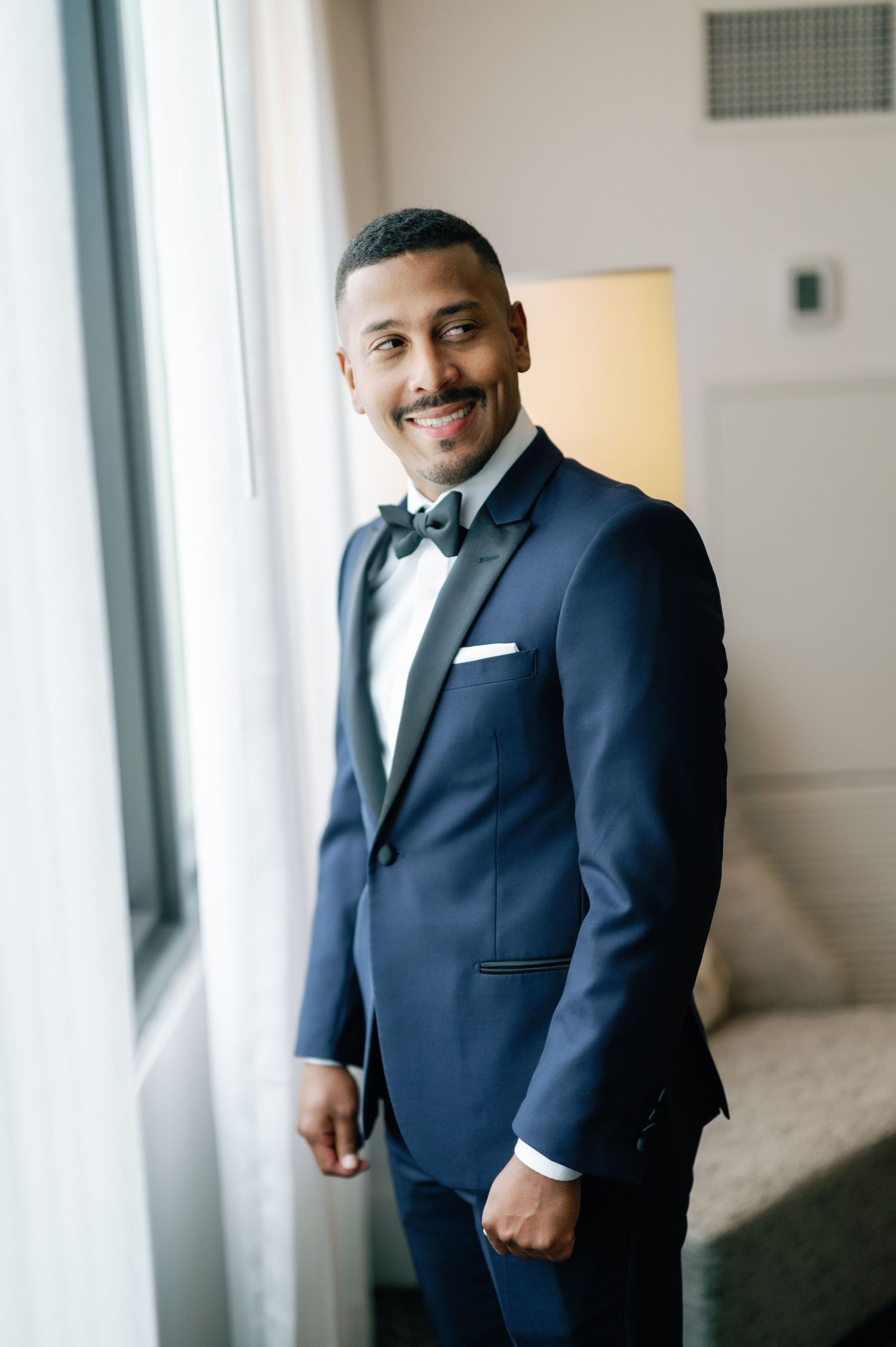 A person wearing a navy blue suit and bow tie poses confidently in a bright hallway setting.