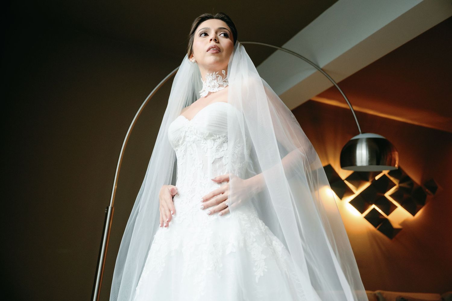 A bride in a flowing white wedding gown and long veil poses dramatically beneath a modern ceiling light fixture.