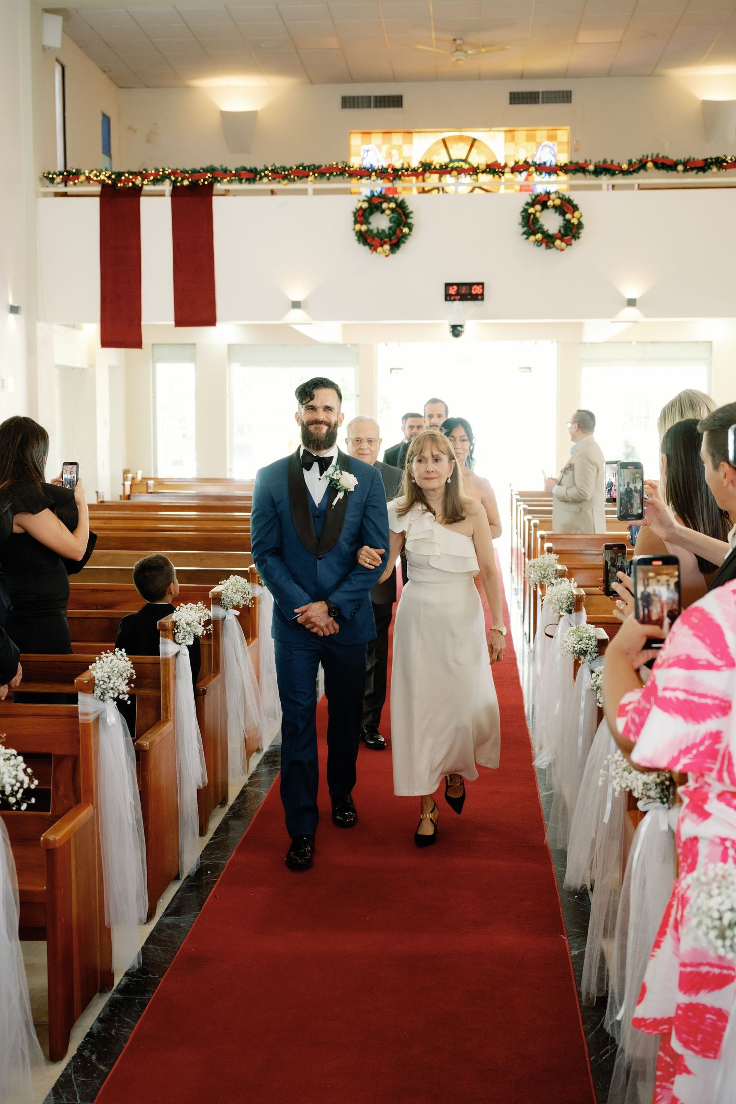 Wedding ceremony in progress showing decorated church aisle with red carpet and Christmas garland along the pews.