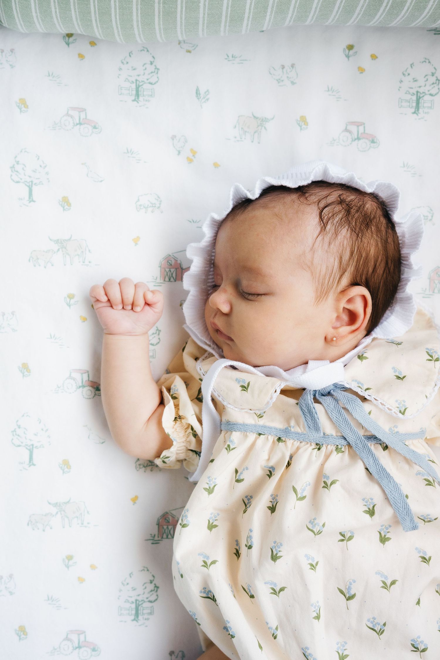 Sleeping newborn baby in floral dress and white bonnet lying in a crib with animal print bedding.