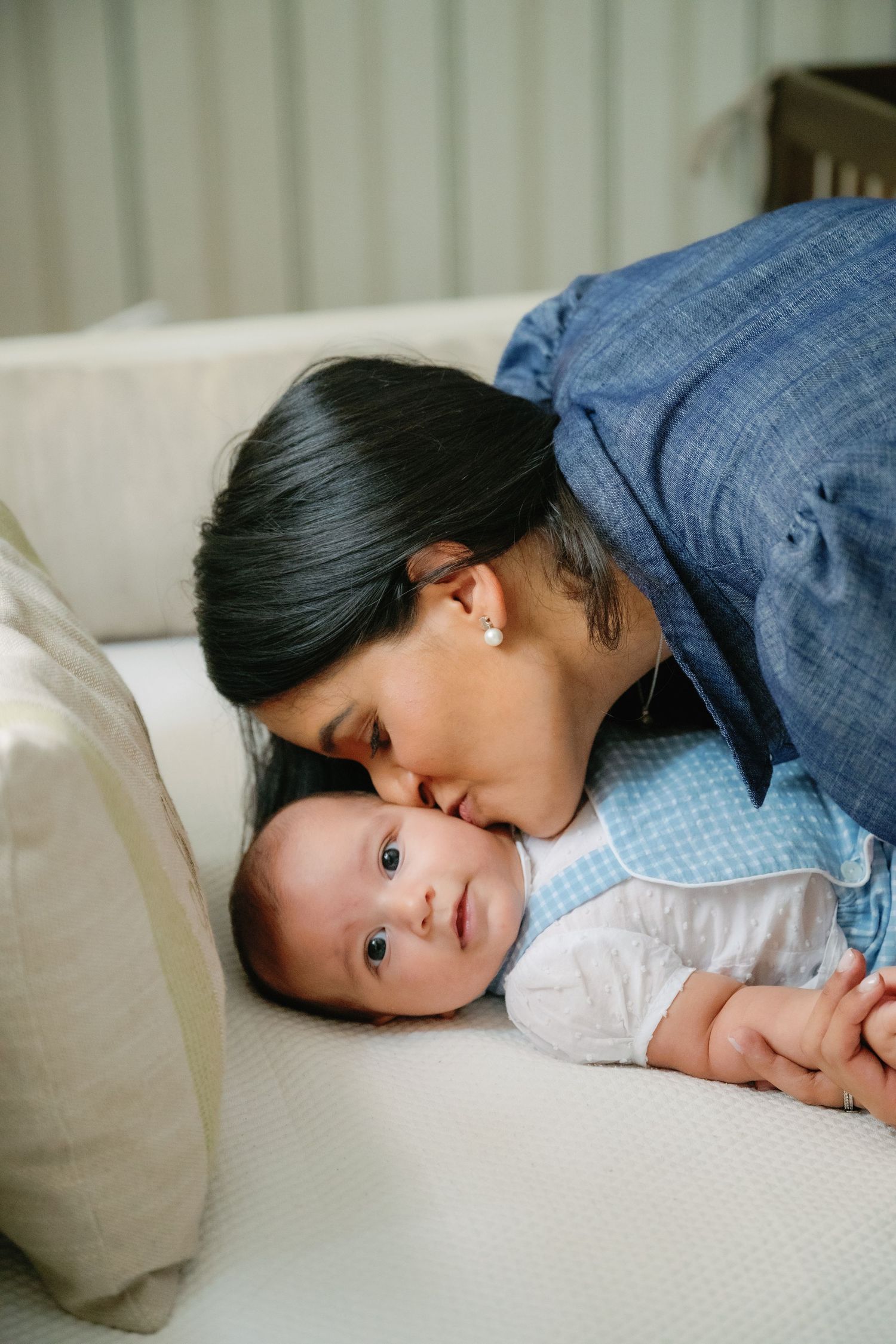 A loving mother gently kisses her newborn baby lying on a white couch, sharing a tender moment.