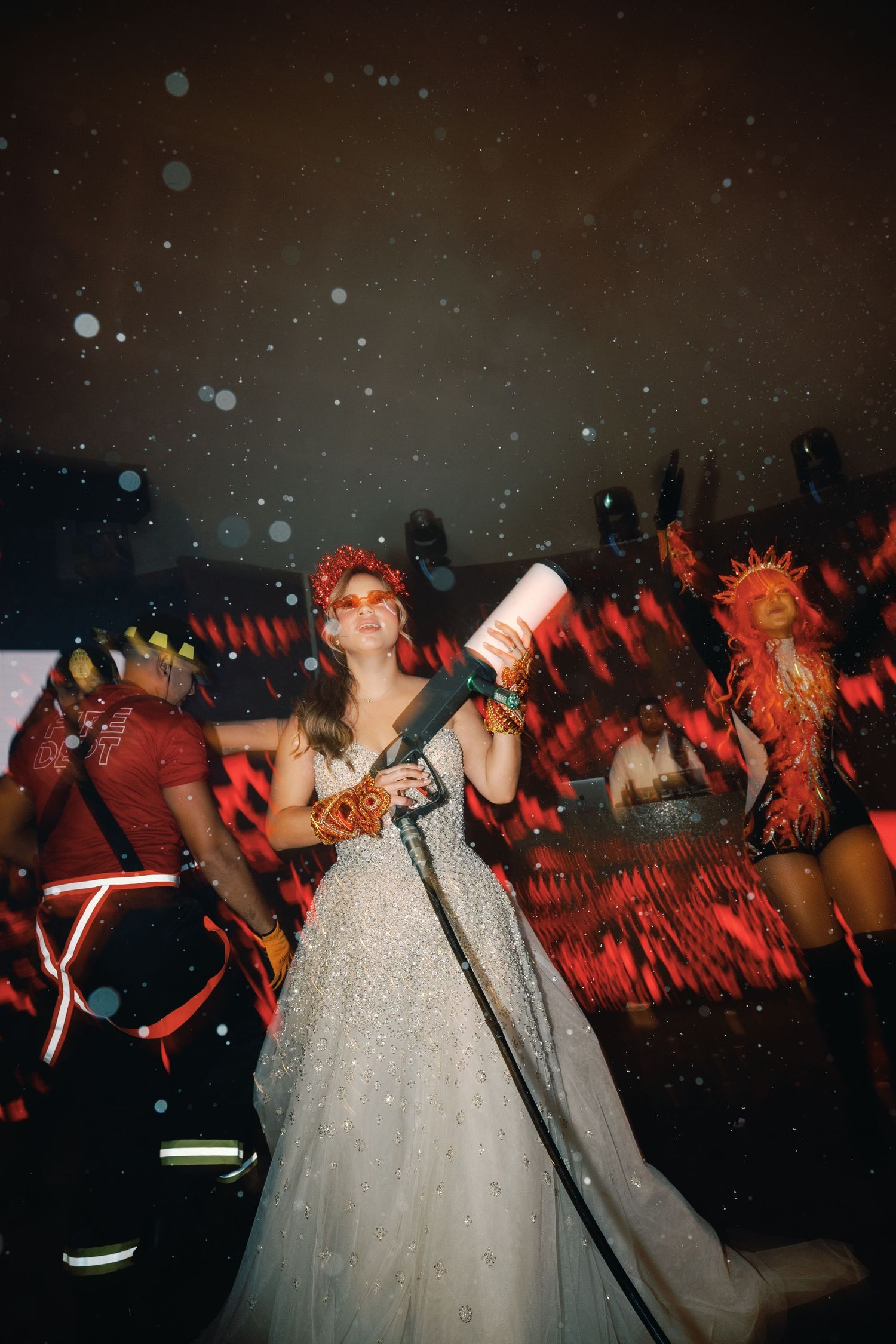 A person in a sparkly white gown holds a drink bottle at a festive party with red lighting and blurred background effects.