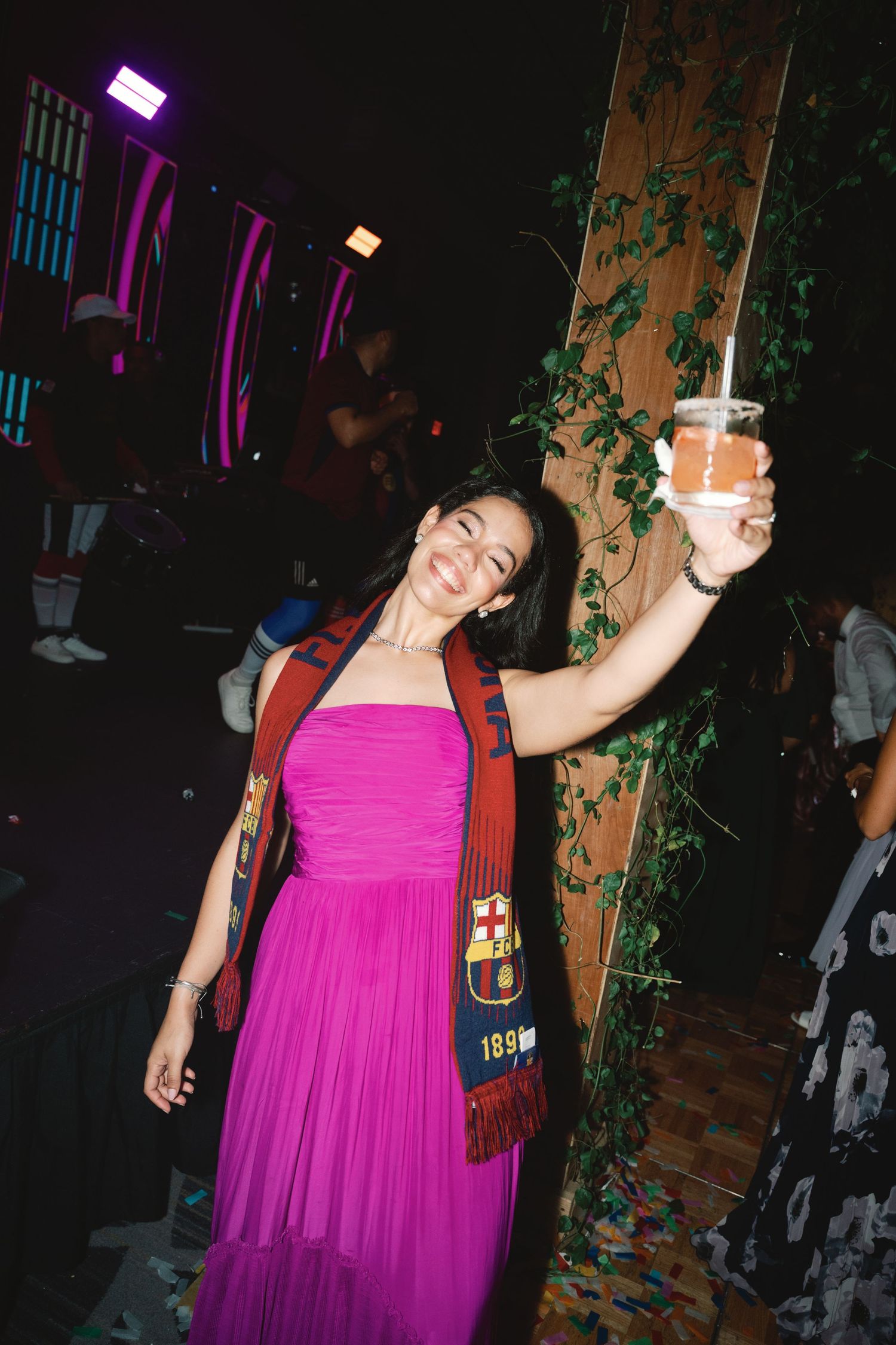 Woman in bright pink dress holds up drink at nightclub party with purple lighting.