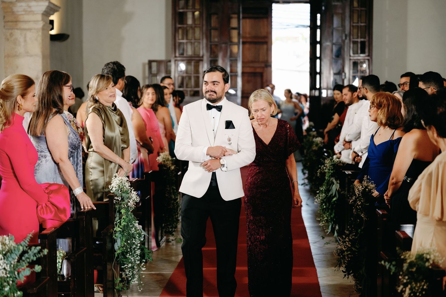 Wedding guests dance and celebrate on an elegant dance floor surrounded by decorative greenery at an indoor reception.