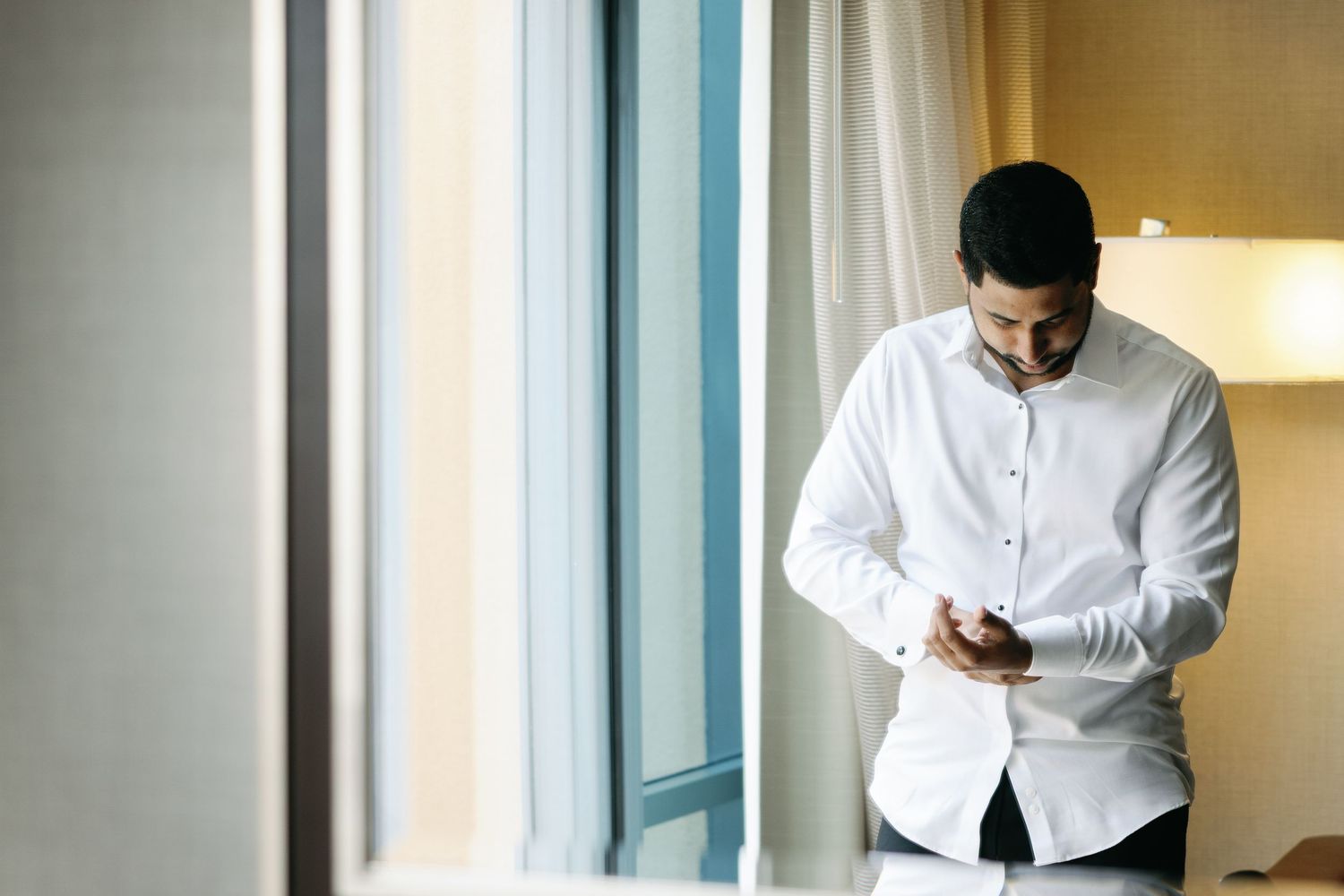 A person in a white button-up shirt stands by a window while looking at their mobile phone.