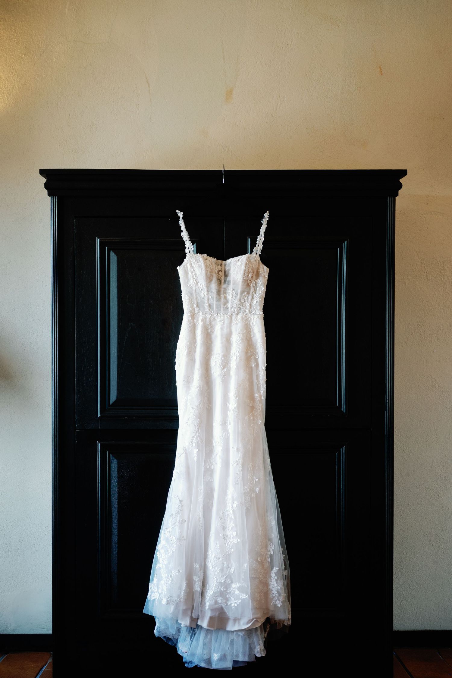 White lace wedding dress hangs elegantly against a dark black wardrobe in dramatic lighting.