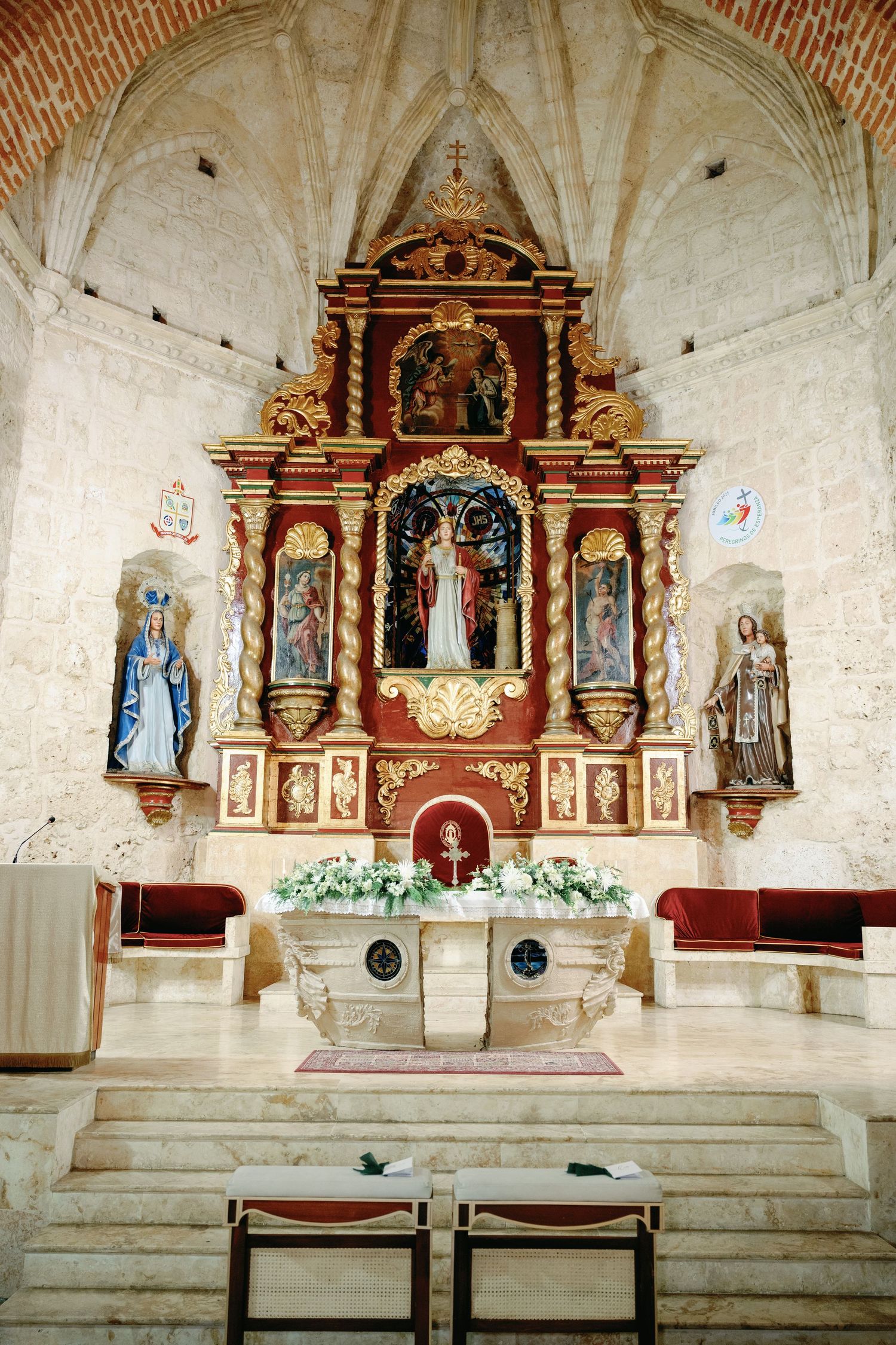 Interior view of a historic church altar with ornate baroque decorations, white stone walls, and religious statues.
