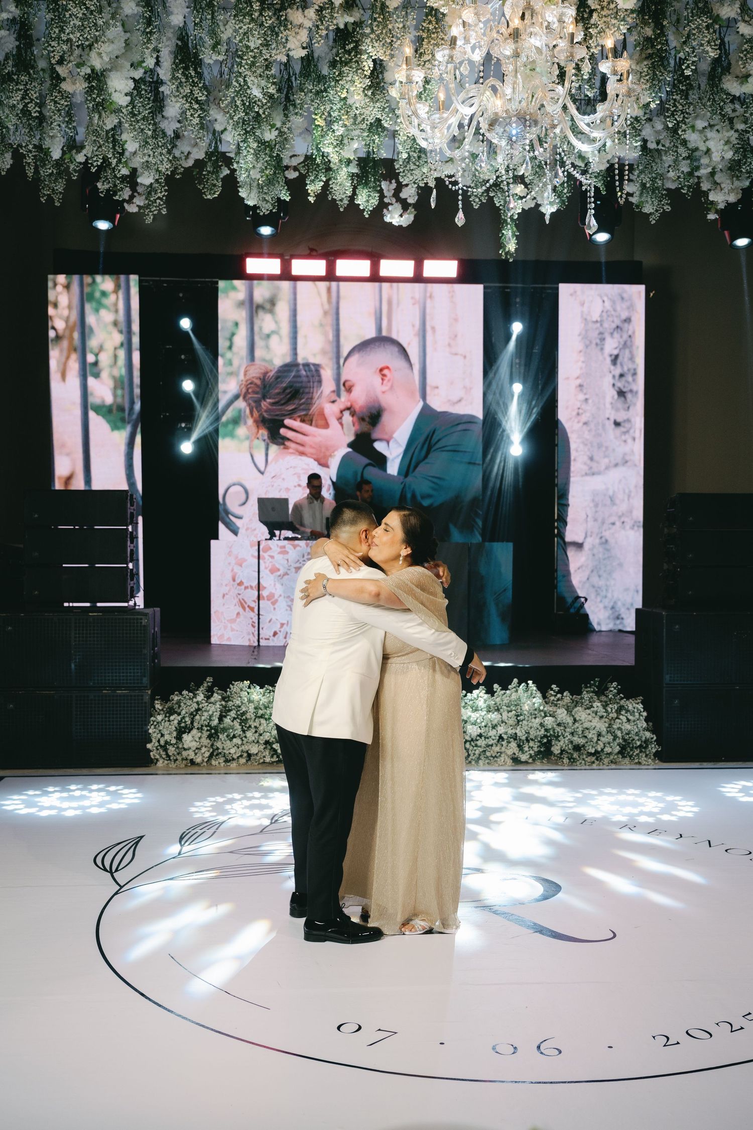 A couple shares a romantic dance on an elegant white dance floor beneath crystal chandeliers and floral decorations.