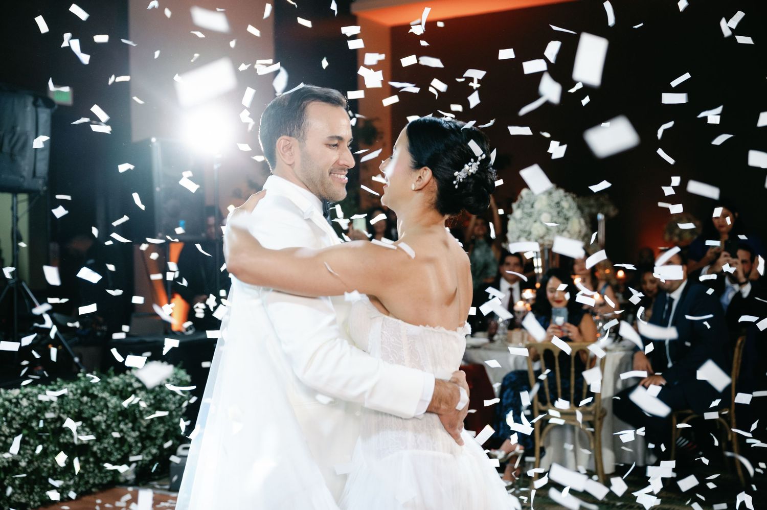 Couple dancing during their first dance at wedding reception with white confetti falling around them.