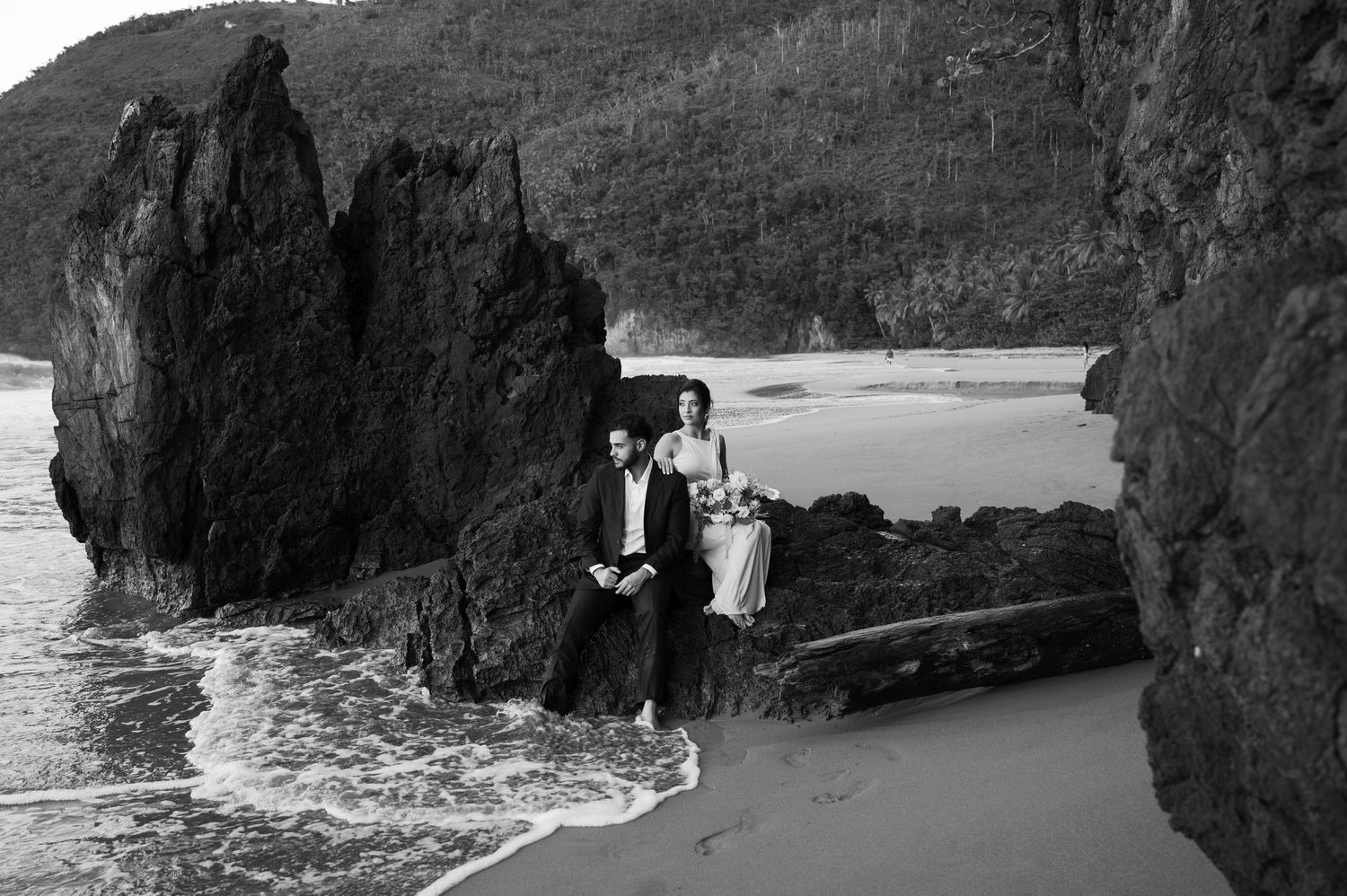 Dramatic black and white coastal wedding photo with rocky cliffs and ocean waves.