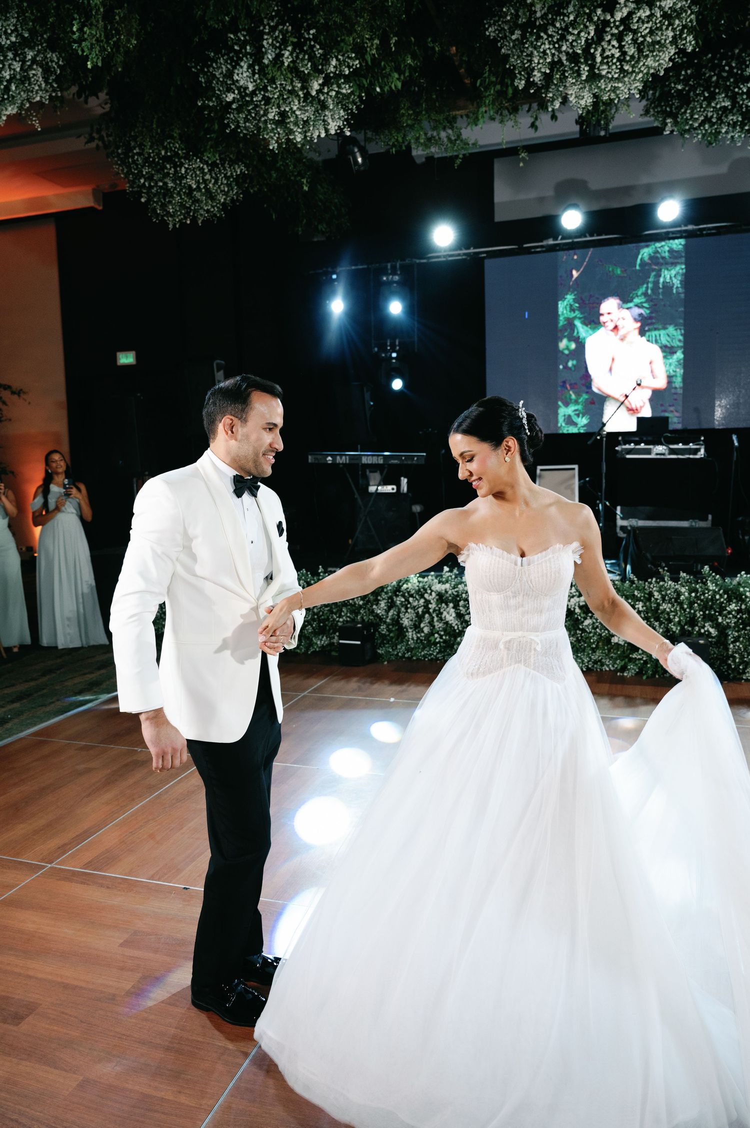 Newlyweds share a romantic dance together on an illuminated dance floor under decorative floral arrangements.