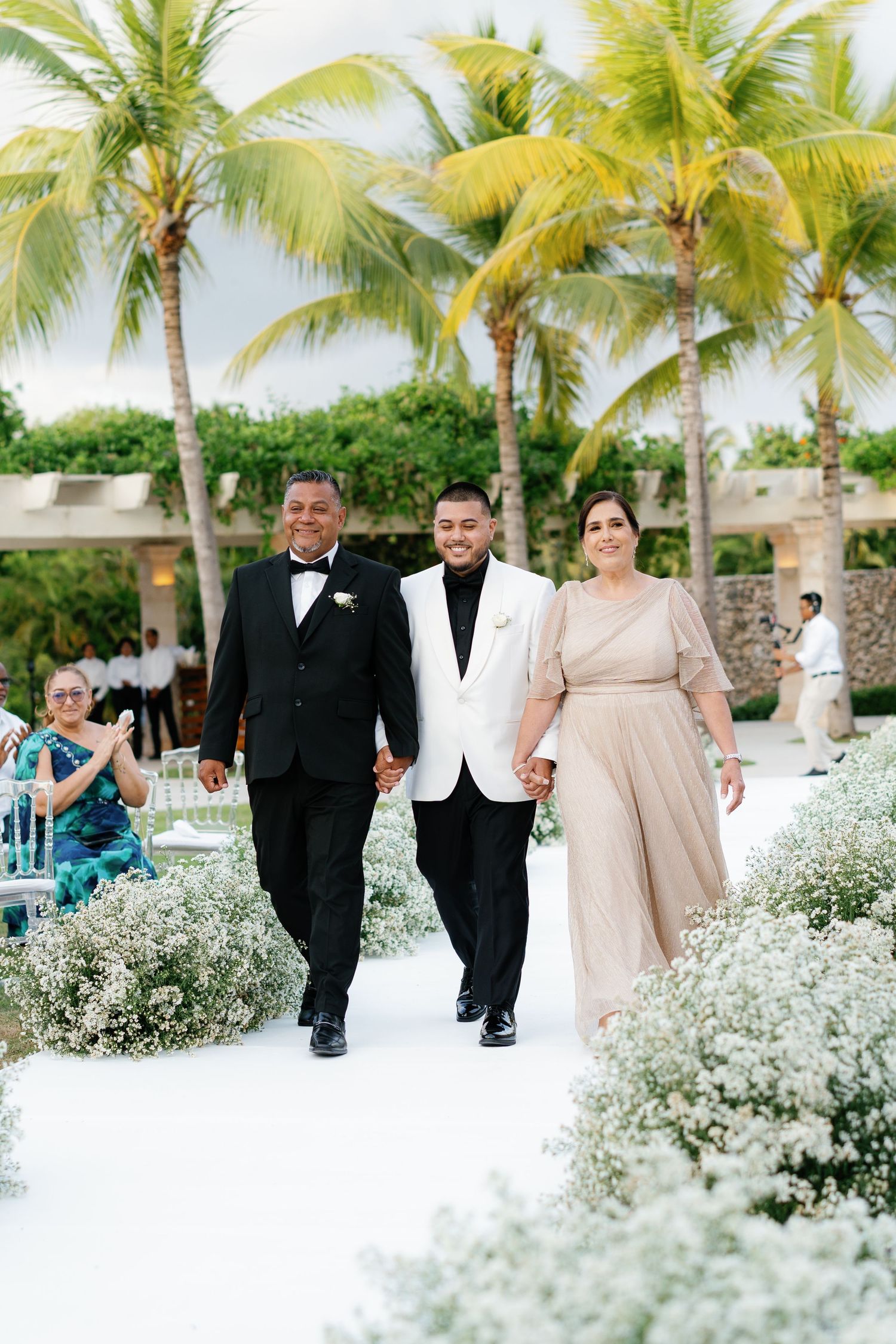 Wedding party walking down a white aisle lined with baby's breath flowers and tropical palm trees.
