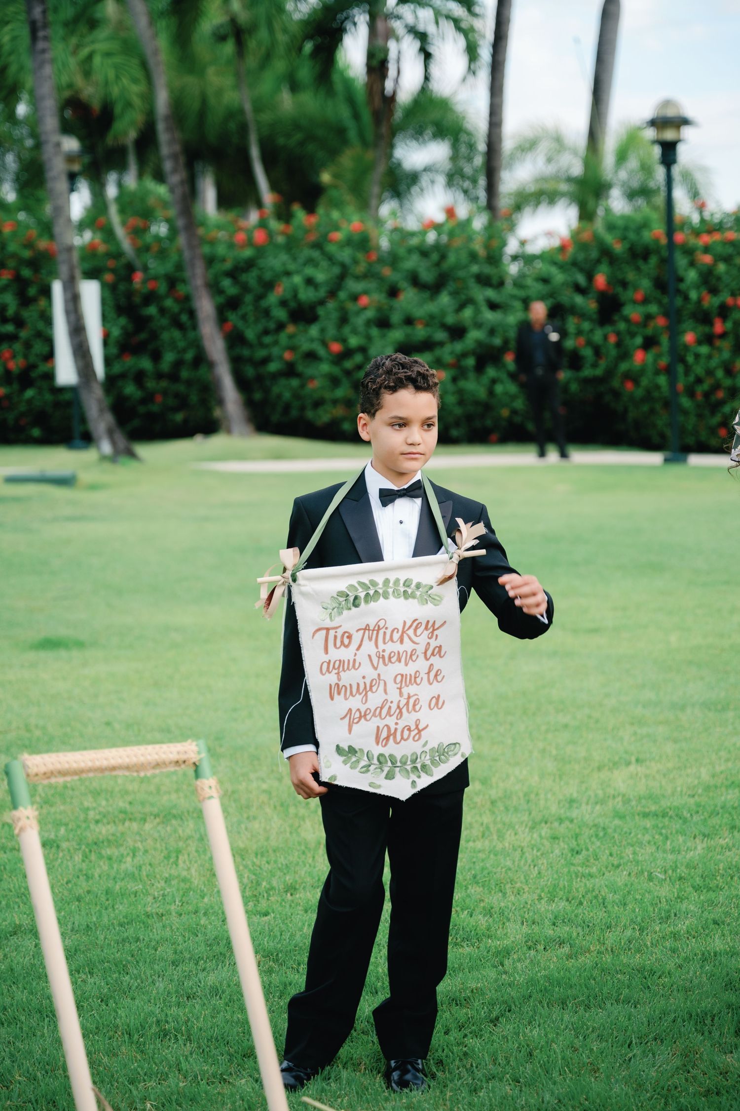 Young ring bearer in a tuxedo holds decorative signs on wooden stands at an outdoor wedding ceremony.