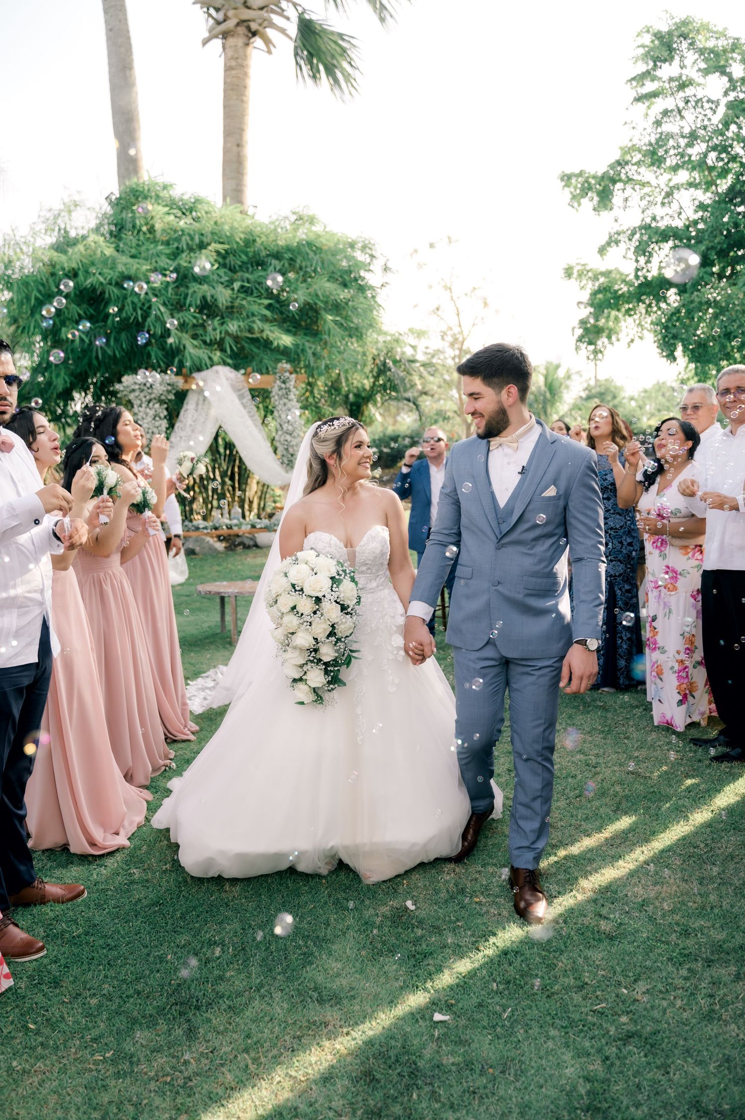 Wedding party celebrates on a grassy lawn with bridesmaids in pink dresses and groomsmen in gray suits.