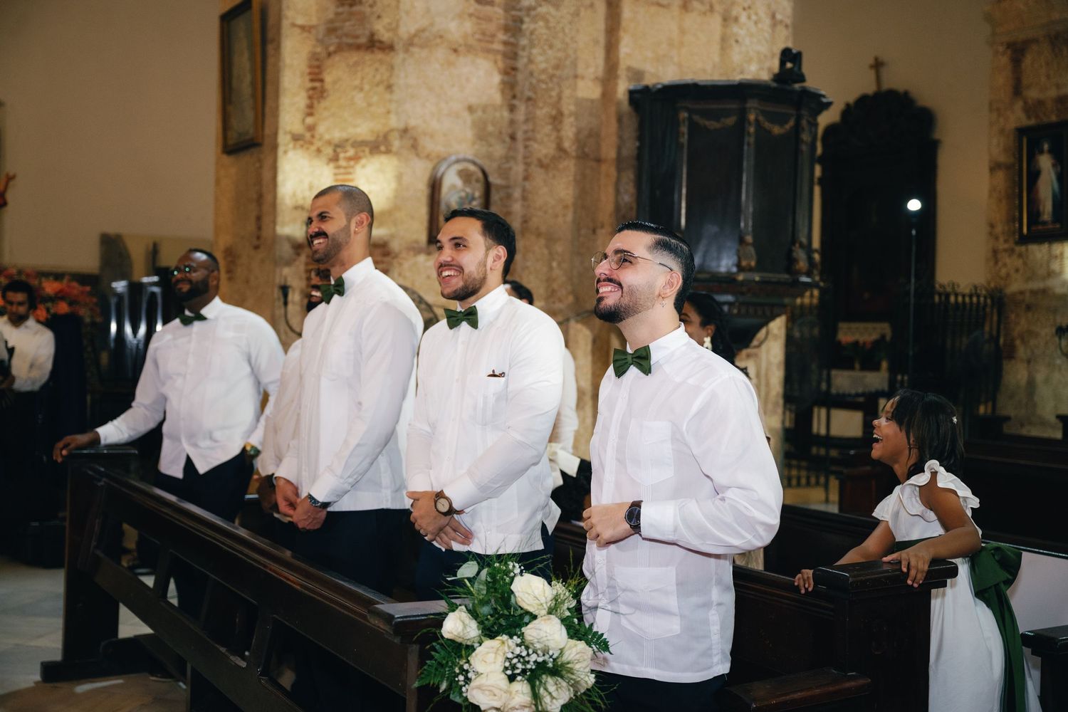 Wedding party members in white dress shirts and formal attire interact in an elegant stone interior setting.