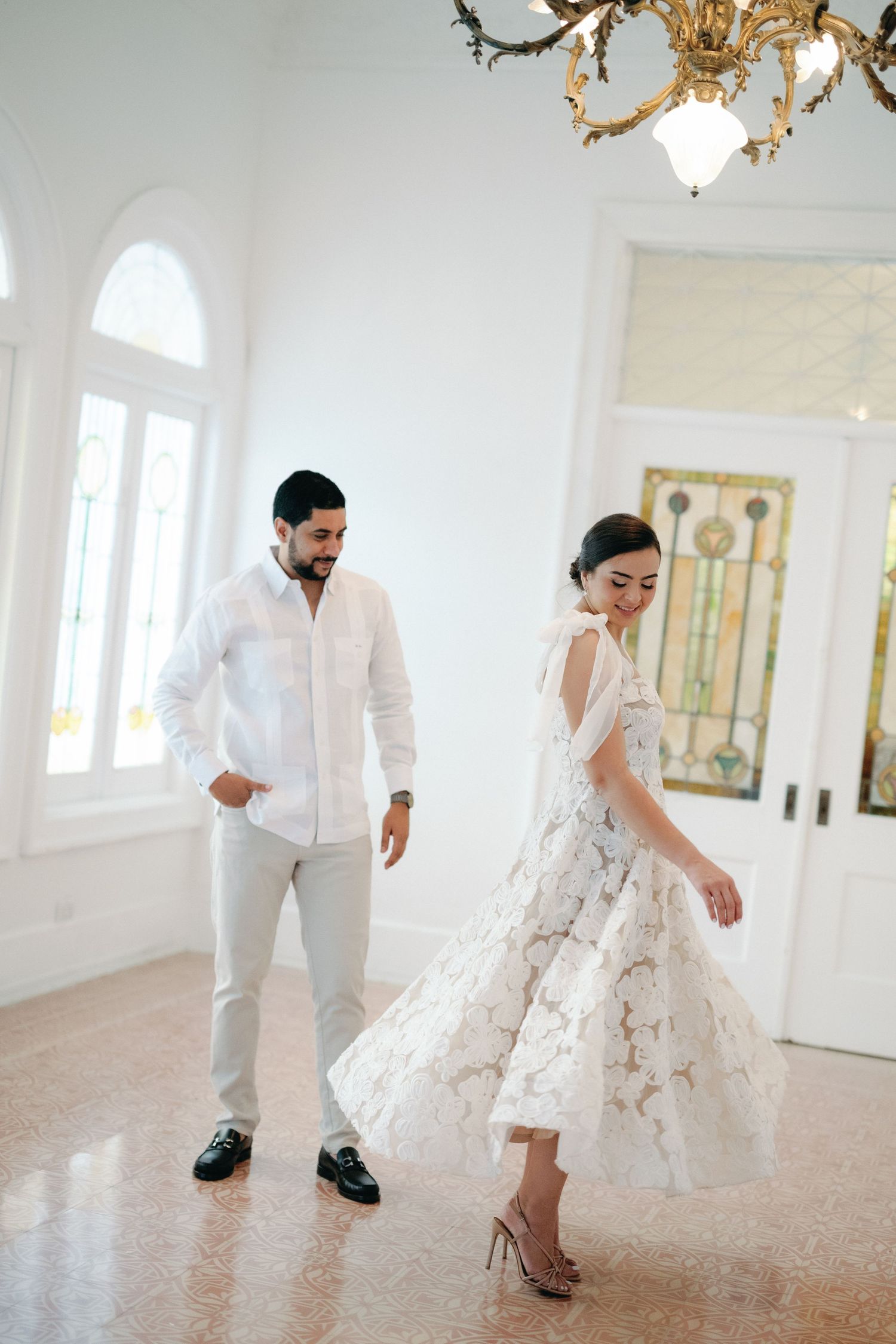 A bride and groom dance playfully in an elegant white room with hardwood floors and natural light streaming through windows.