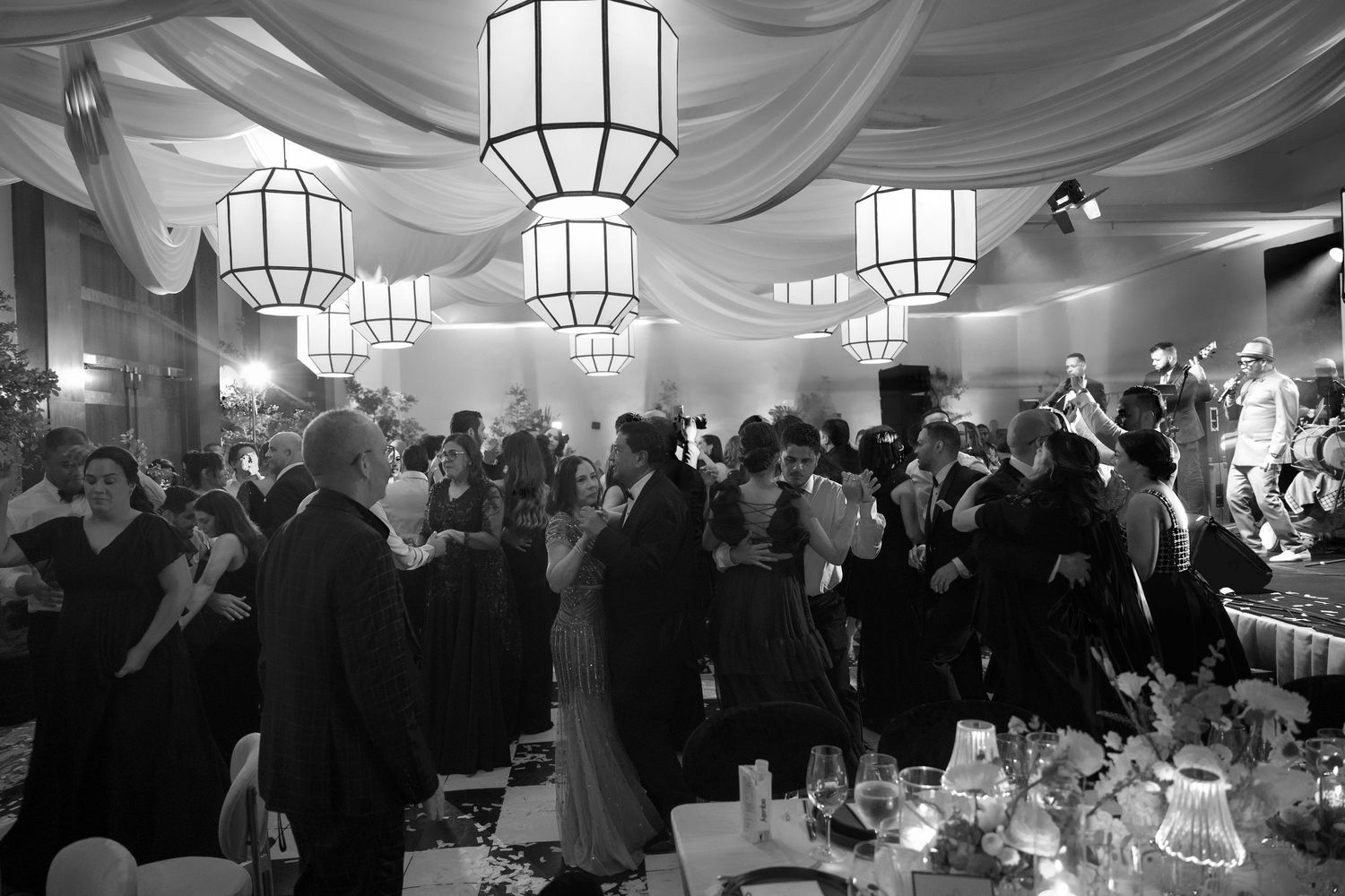 A crowded black and white reception with elegant hanging lanterns and draped fabric decor illuminating the formal gathering.
