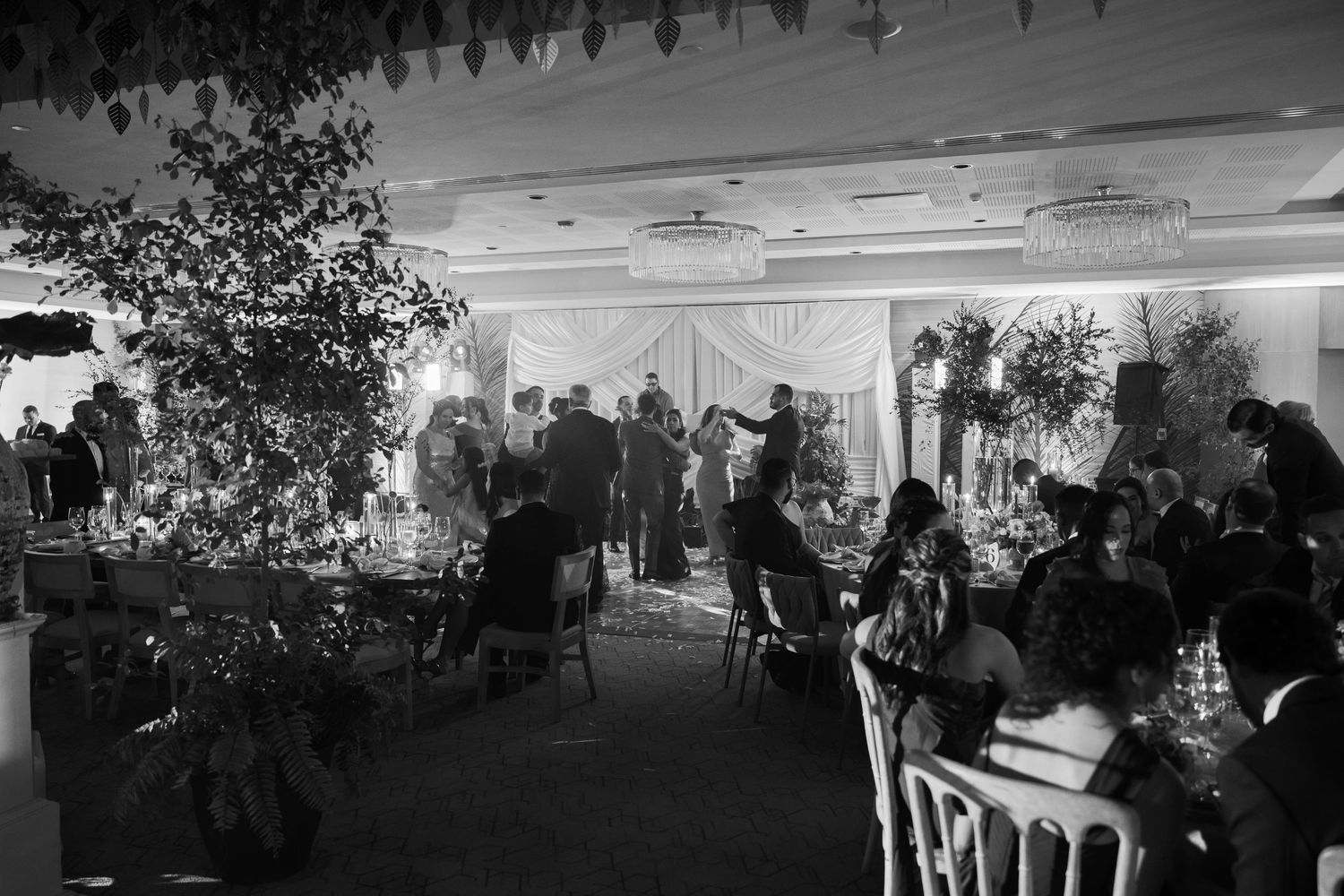 Black and white photo of an elegant indoor event with guests seated at tables among decorative floral arrangements.