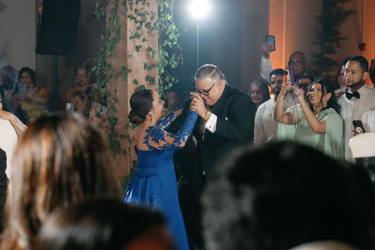 A couple shares a dance at an elegant wedding reception with guests watching and a bright light illuminating the scene.