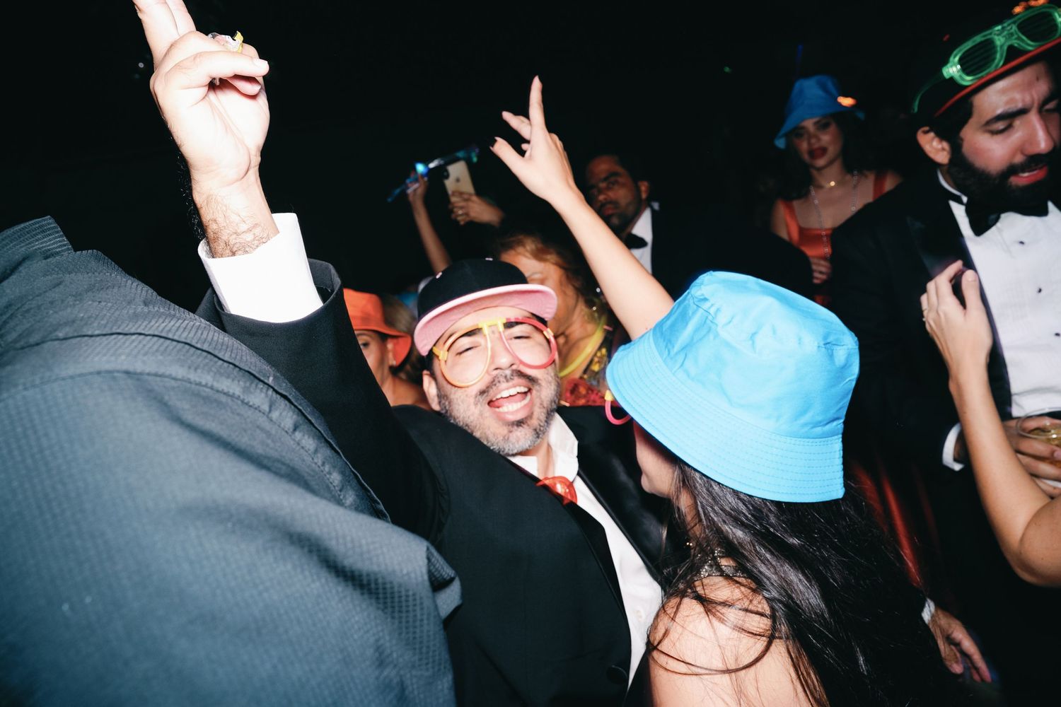 Party goers dance and celebrate at a nighttime event wearing colorful hats and festive attire.