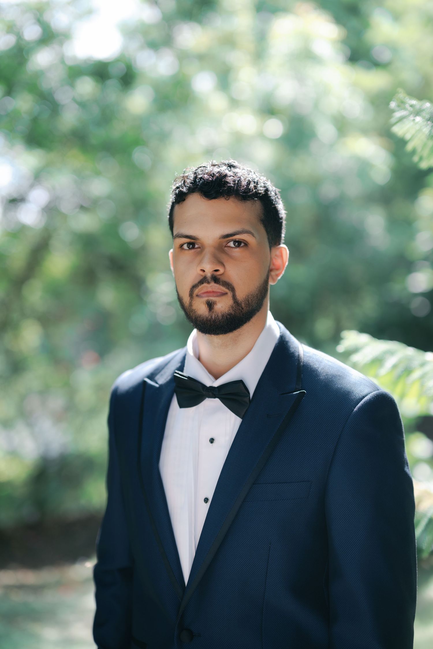 A groom in a classic black tuxedo and bowtie poses outdoors with a natural blurred background.