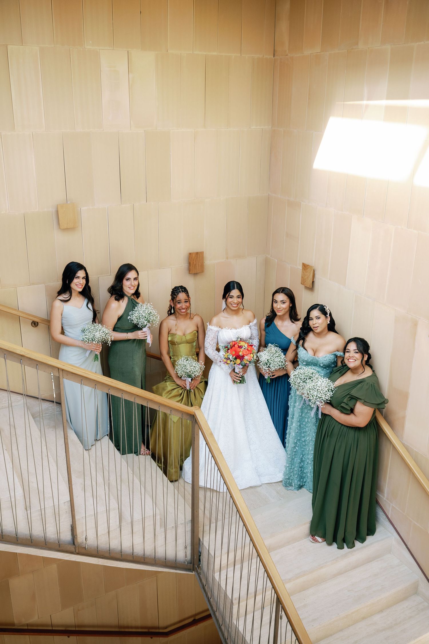 A bridal party in varying shades of green and blue dresses poses elegantly on a light-colored staircase.