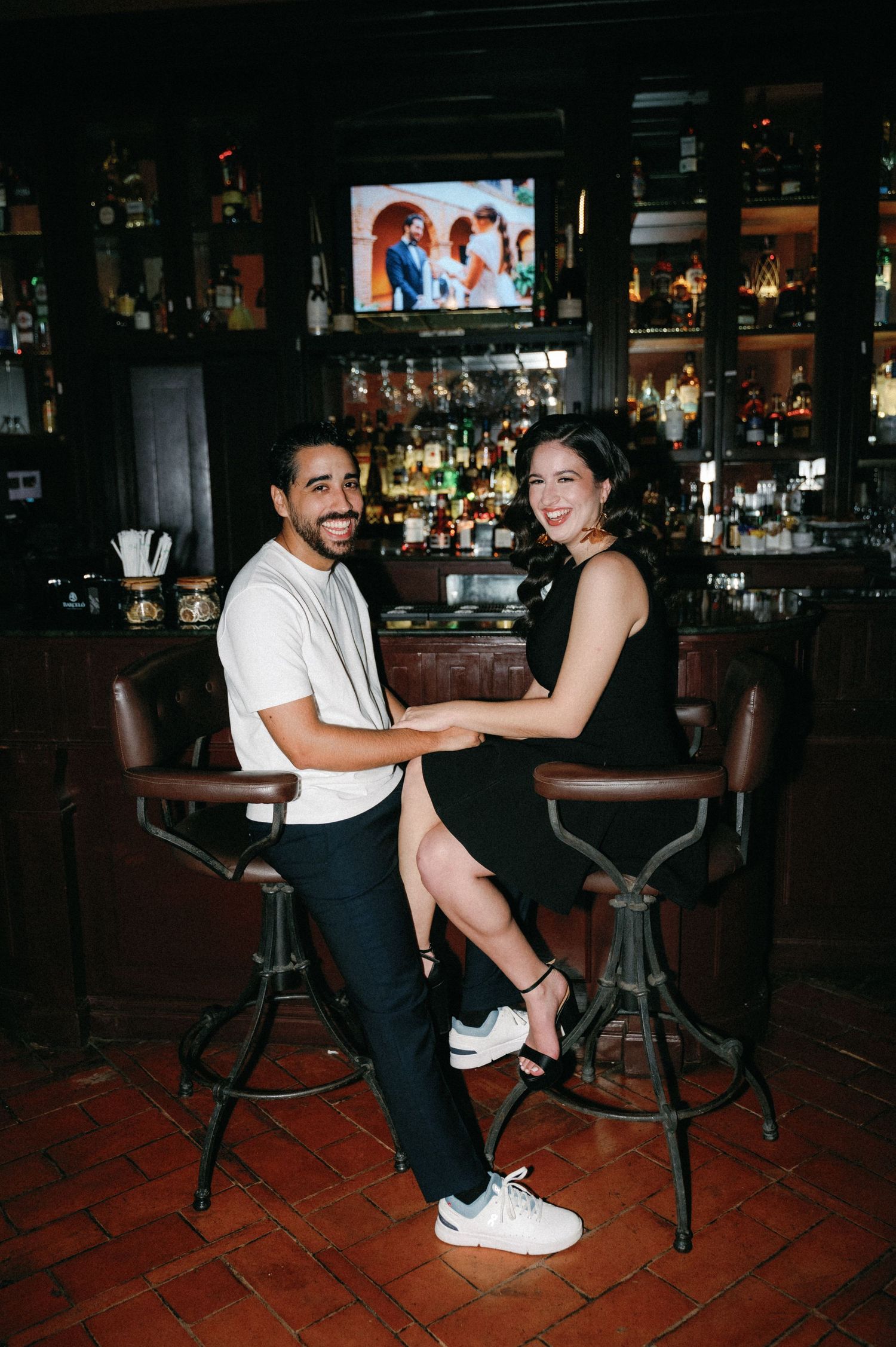 A couple dancing and laughing together at a dimly lit bar with stools and bottles in the background.