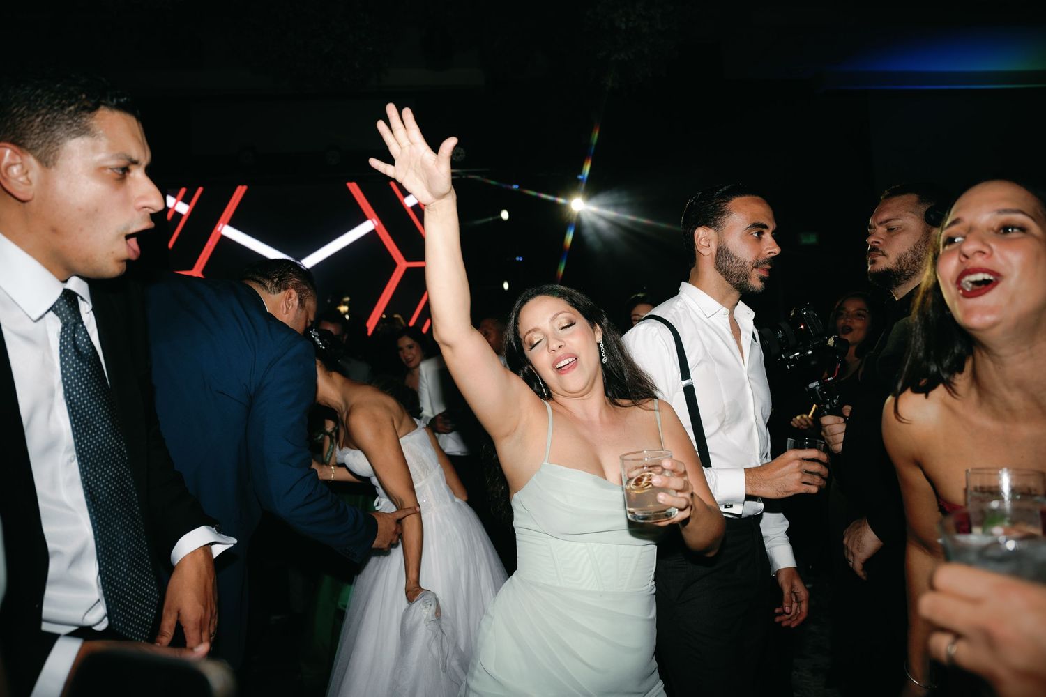 Wedding reception guests dancing and celebrating on a dark dance floor with drinks and festive energy.