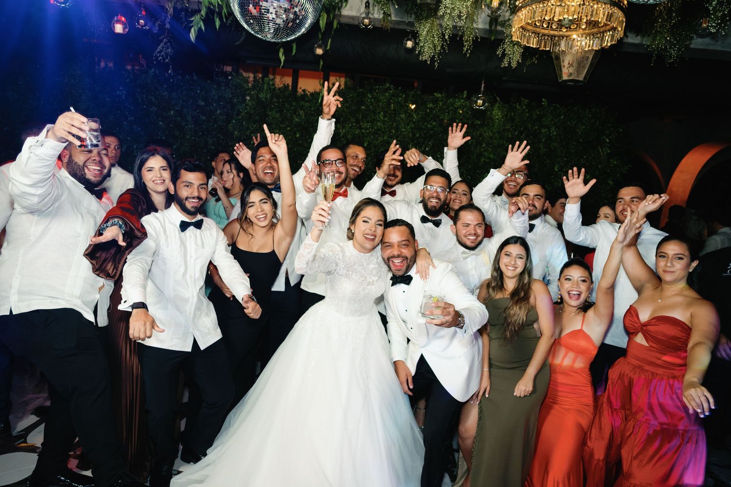 Large group of wedding guests and celebrants posing together with raised arms in celebration at a reception venue.