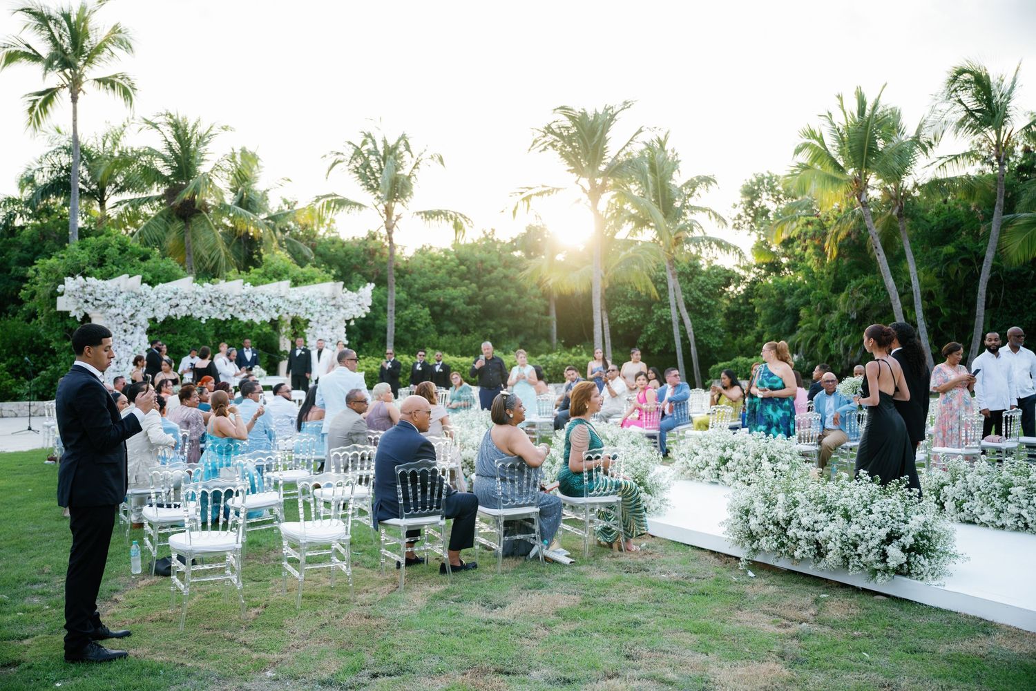 Guests seated at an elegant outdoor wedding ceremony with white chairs and tropical palm trees in the background.