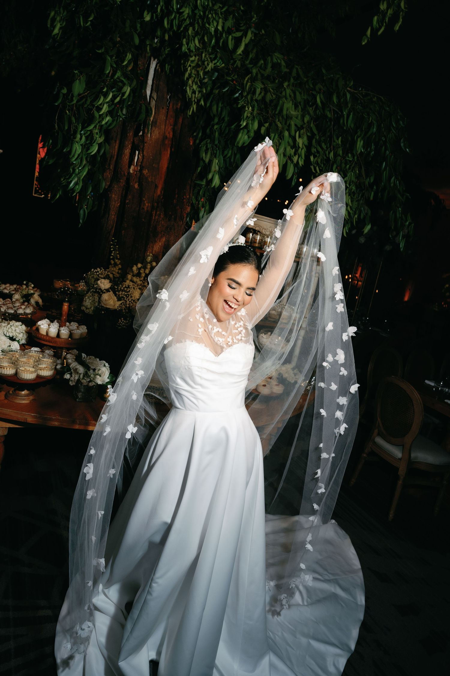 A bride in a white wedding dress playfully poses with her flowing veil in a romantic outdoor setting.