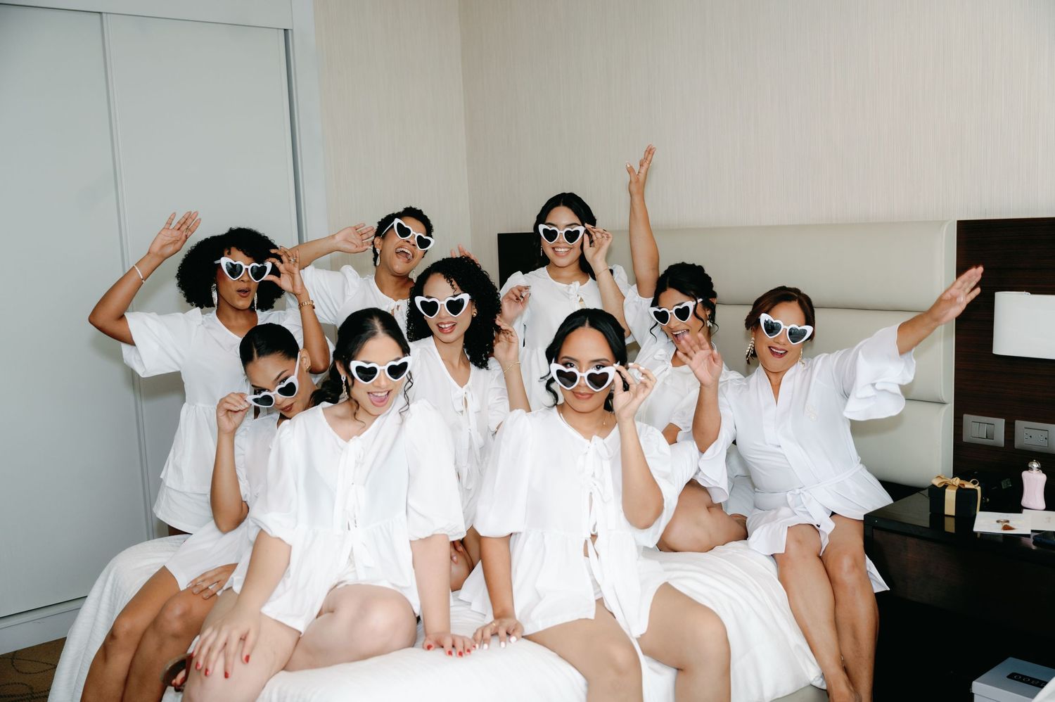 A group in matching white outfits and sunglasses poses playfully on a bed during wedding preparations.