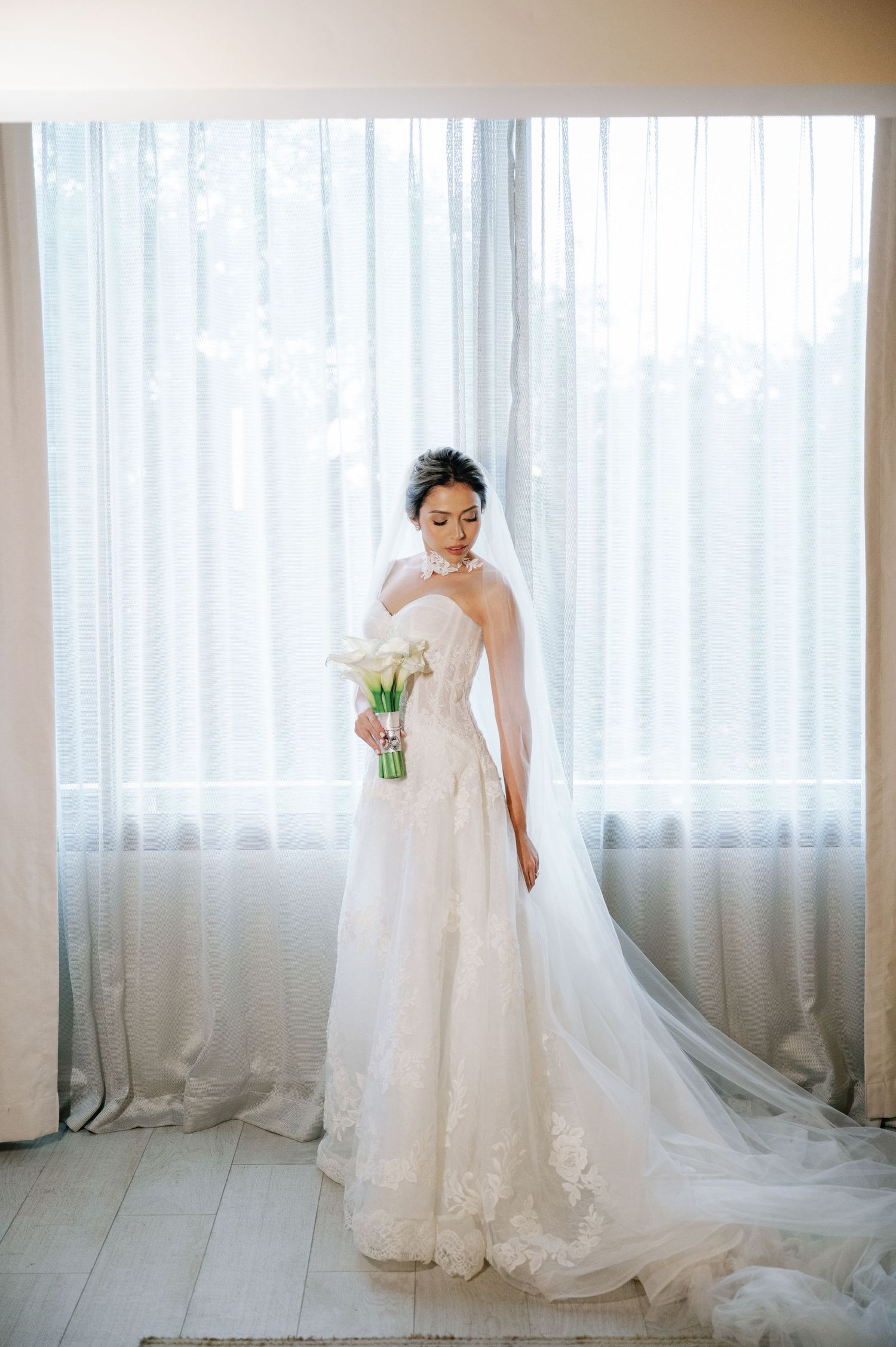 Bride in elegant lace wedding gown with flowing veil poses by window in natural light.