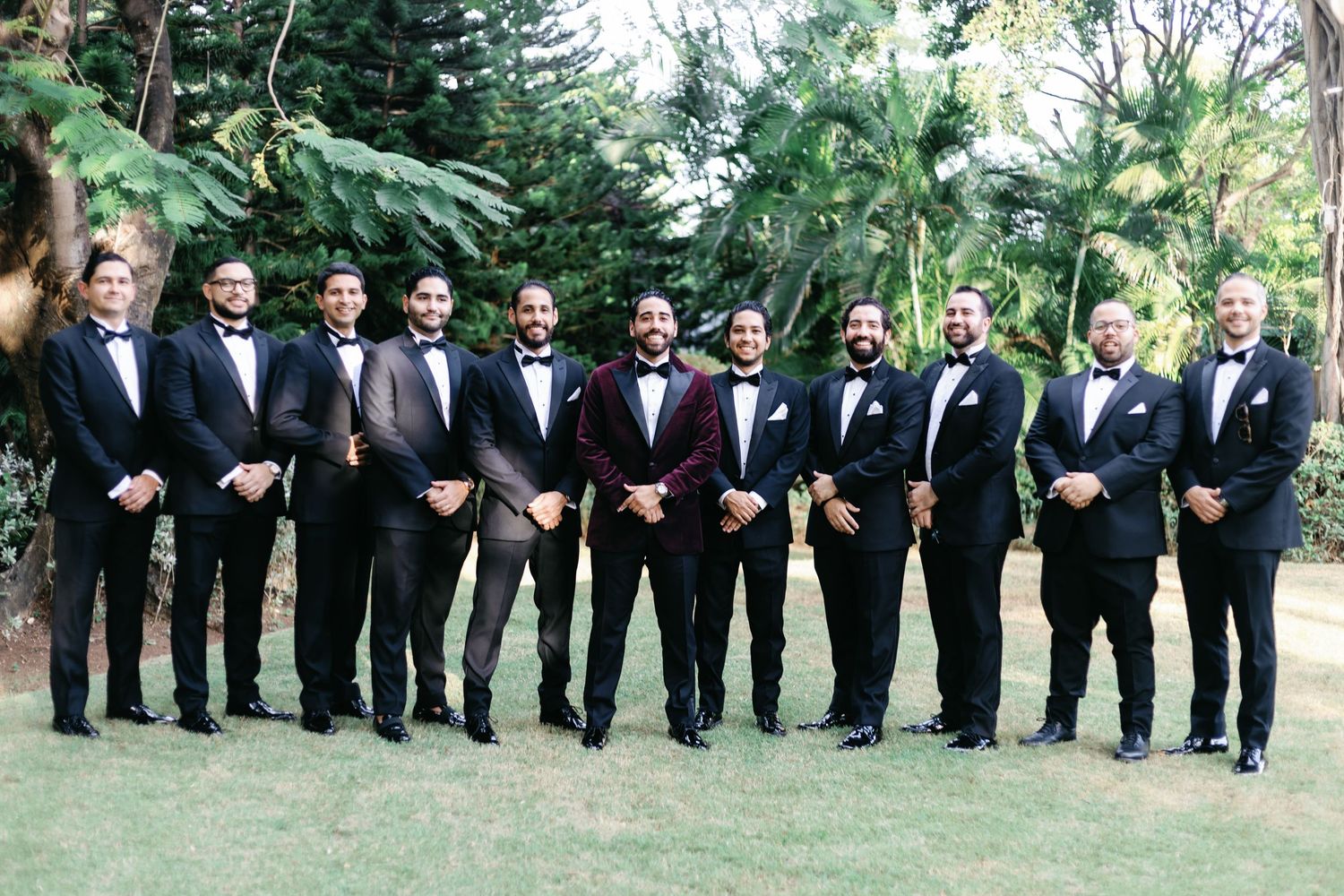 Groomsmen in black tuxedos pose together on a lawn with green trees in the background.
