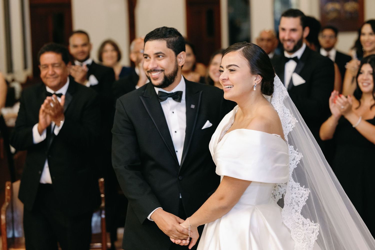 A bride and groom share a joyful moment during their wedding ceremony as guests applaud in the background.