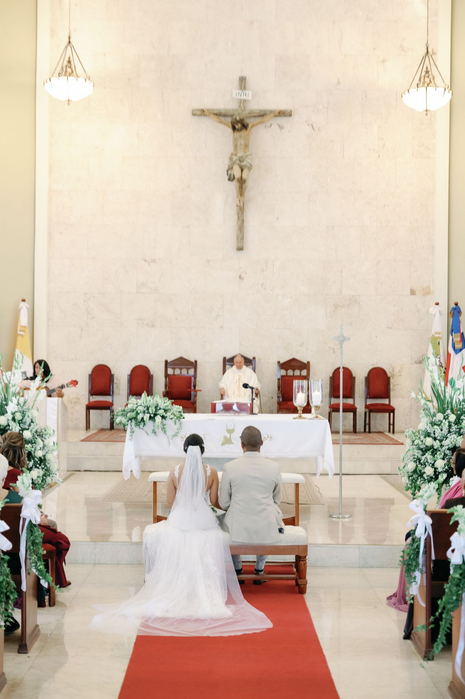 Catholic church interior decorated for a wedding ceremony with red carpet aisle and white floral arrangements.