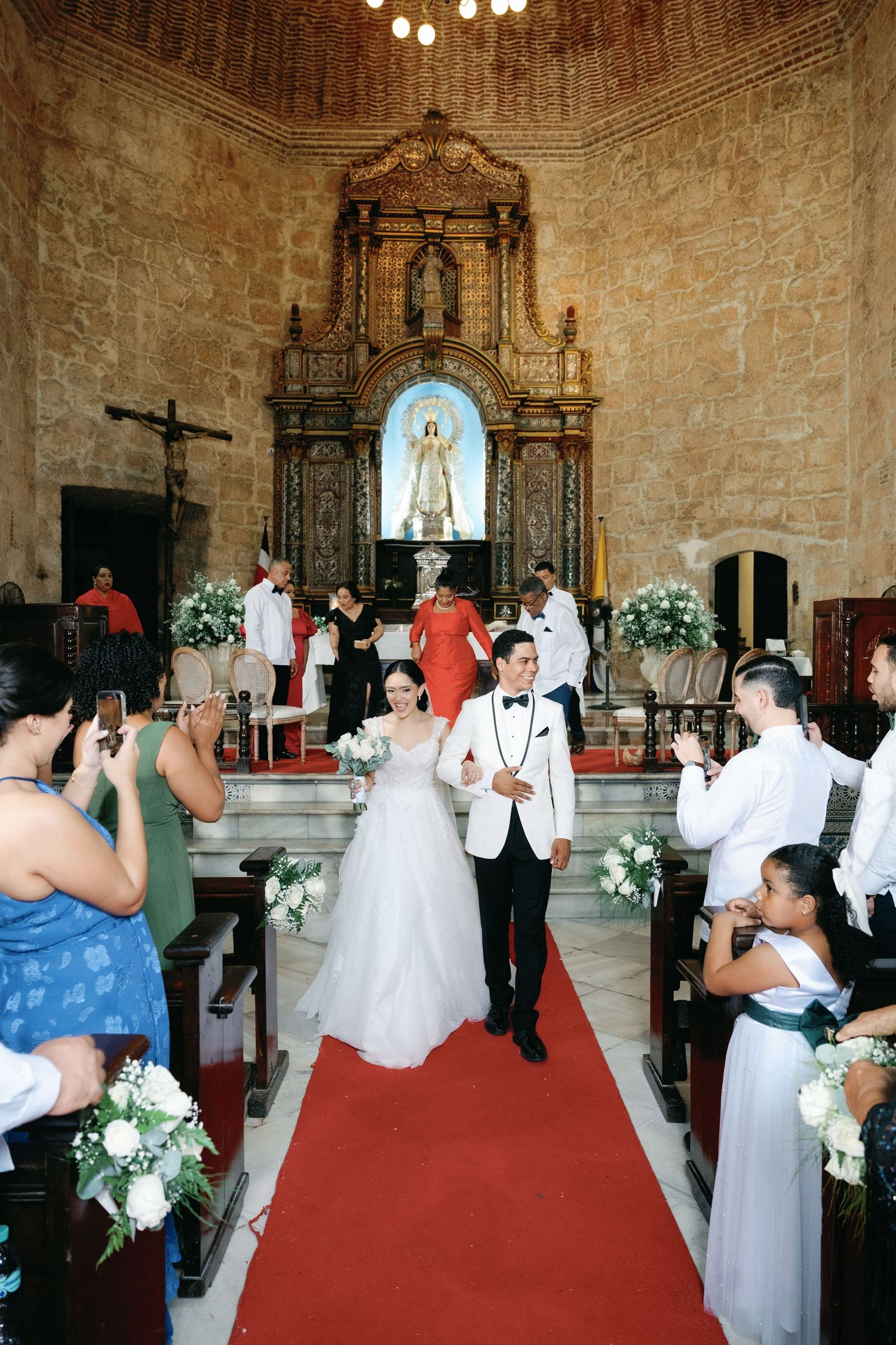 A wedding ceremony taking place in an ornate church with stone walls and a red carpet aisle.