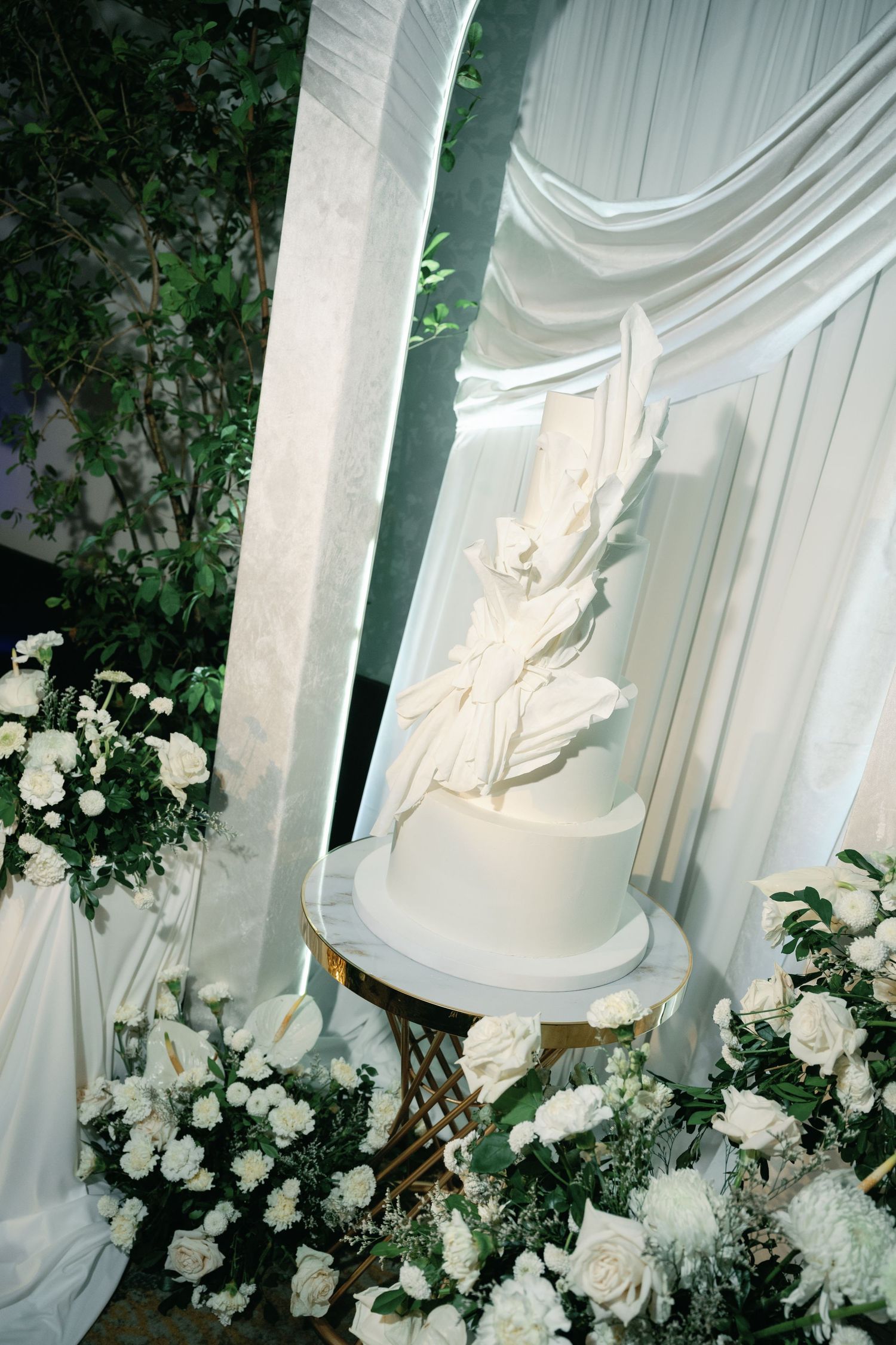 Elegant white wedding cake with delicate sugar flowers surrounded by fresh white roses and greenery against draped fabric.