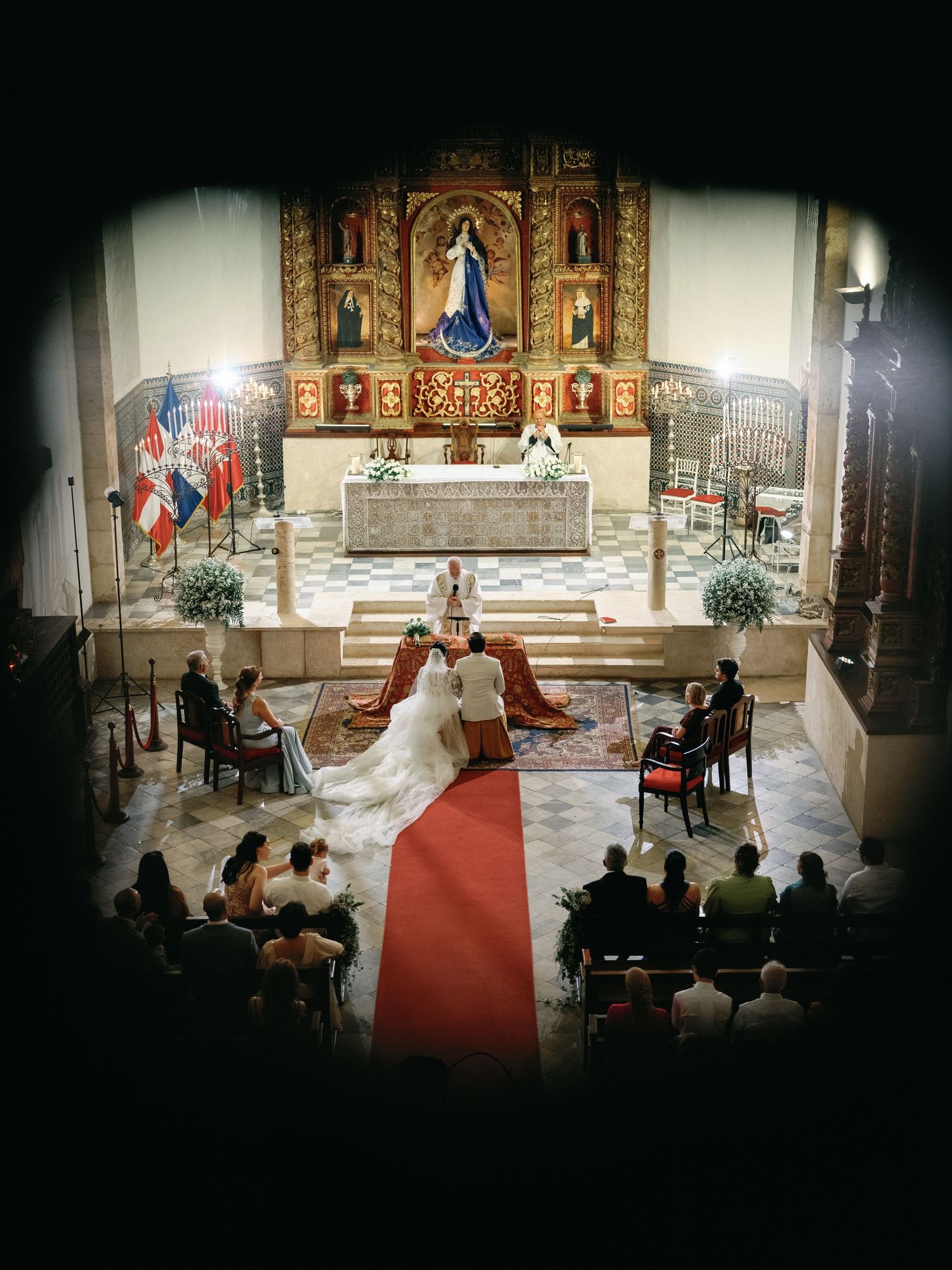 Wedding ceremony taking place in a historic church with ornate altar and red carpet aisle.
