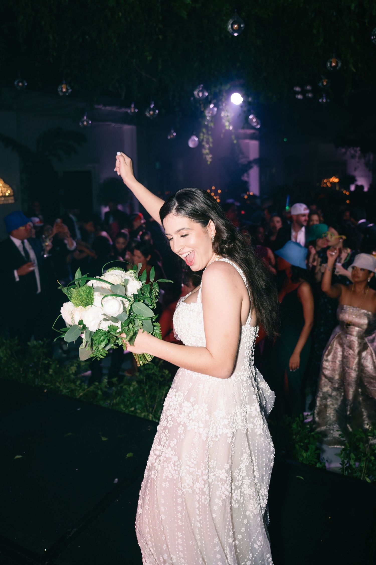 A bride in a white sequined dress holds a bouquet while dancing at a dimly lit wedding reception.