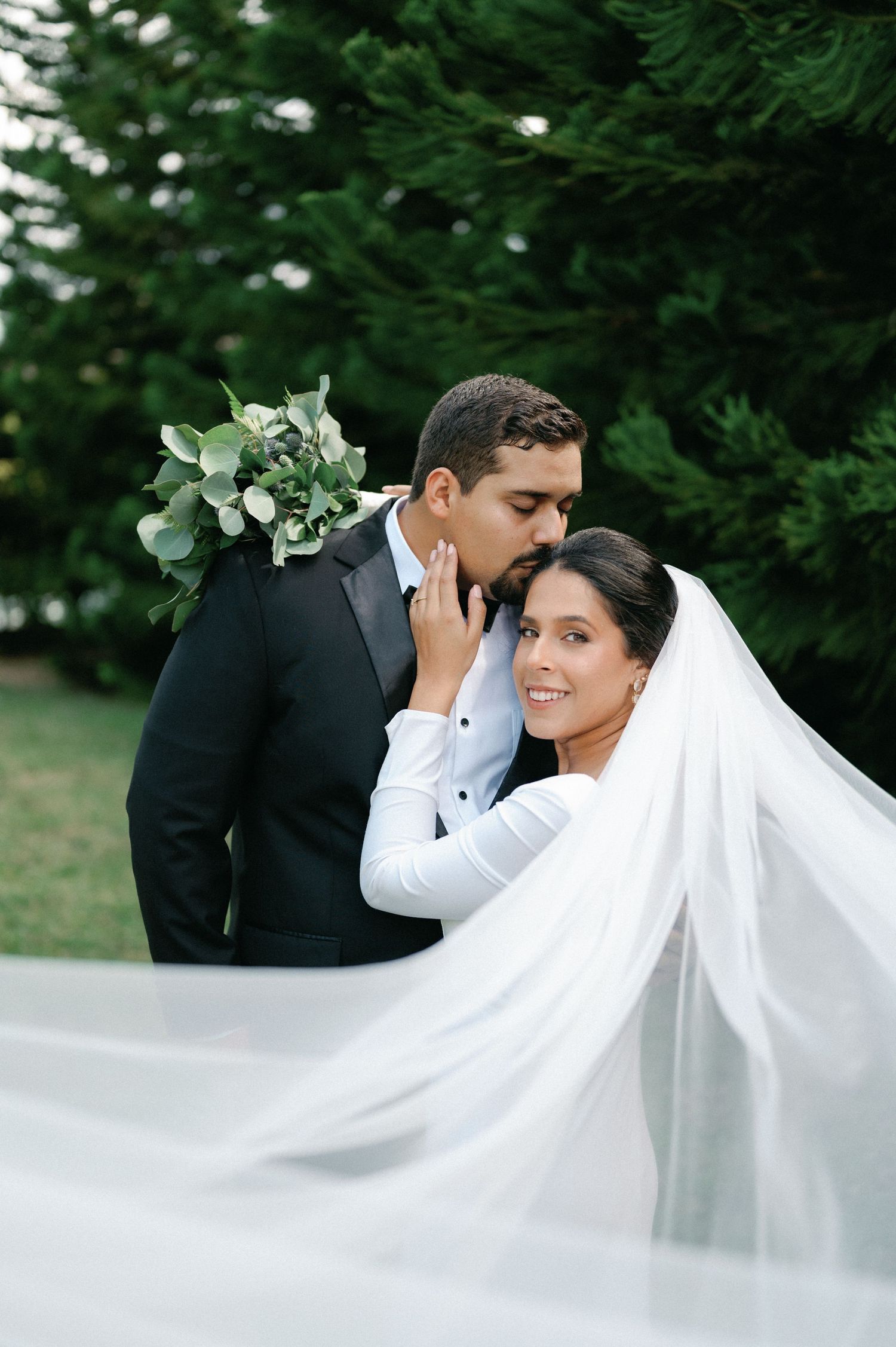 Romantic wedding moment captured through flowing white veil with evergreen trees in background.