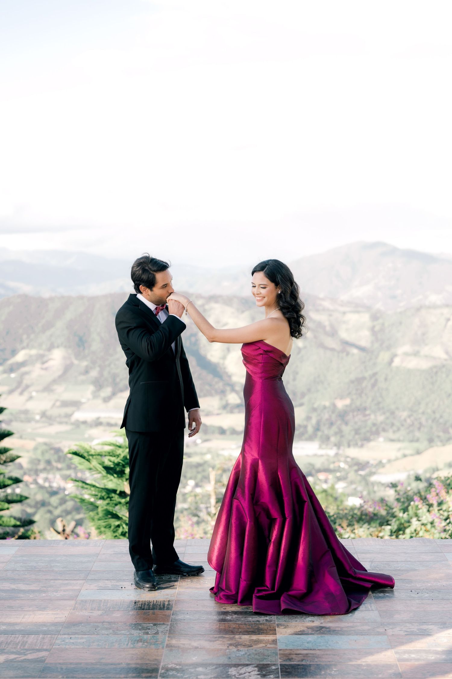 A couple in formal attire share a romantic moment on a mountain overlook, with her wearing a stunning burgundy gown.