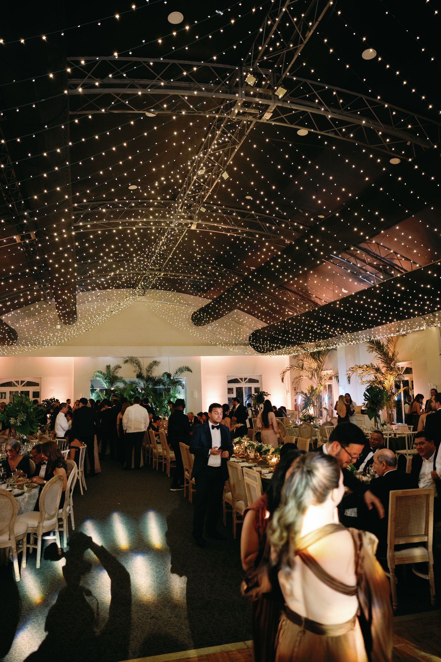 Wedding reception venue with twinkling string lights overhead and guests mingling and dancing on the dance floor.