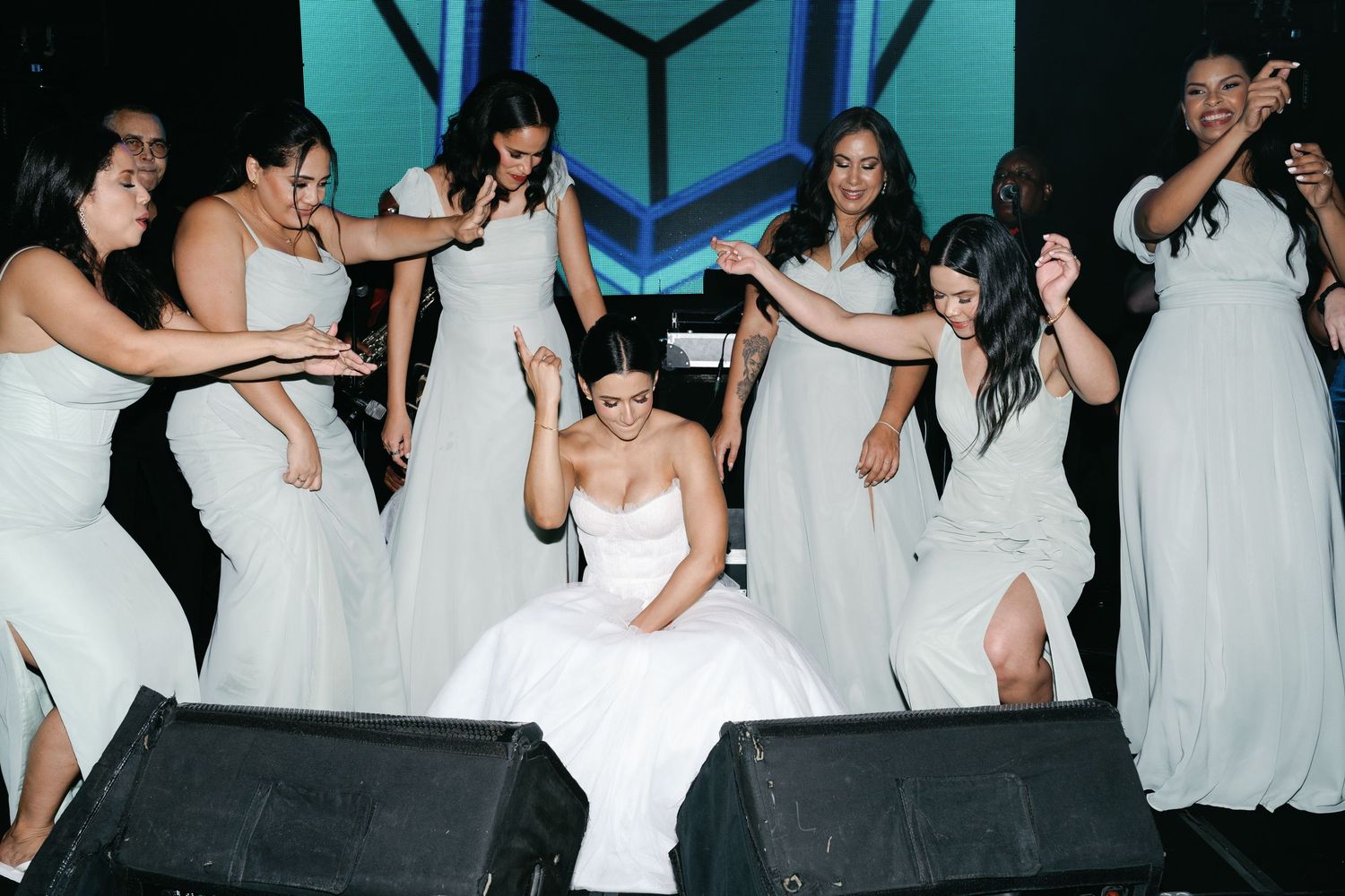 A group of people wearing white dresses dance and celebrate together in a turquoise-colored room.