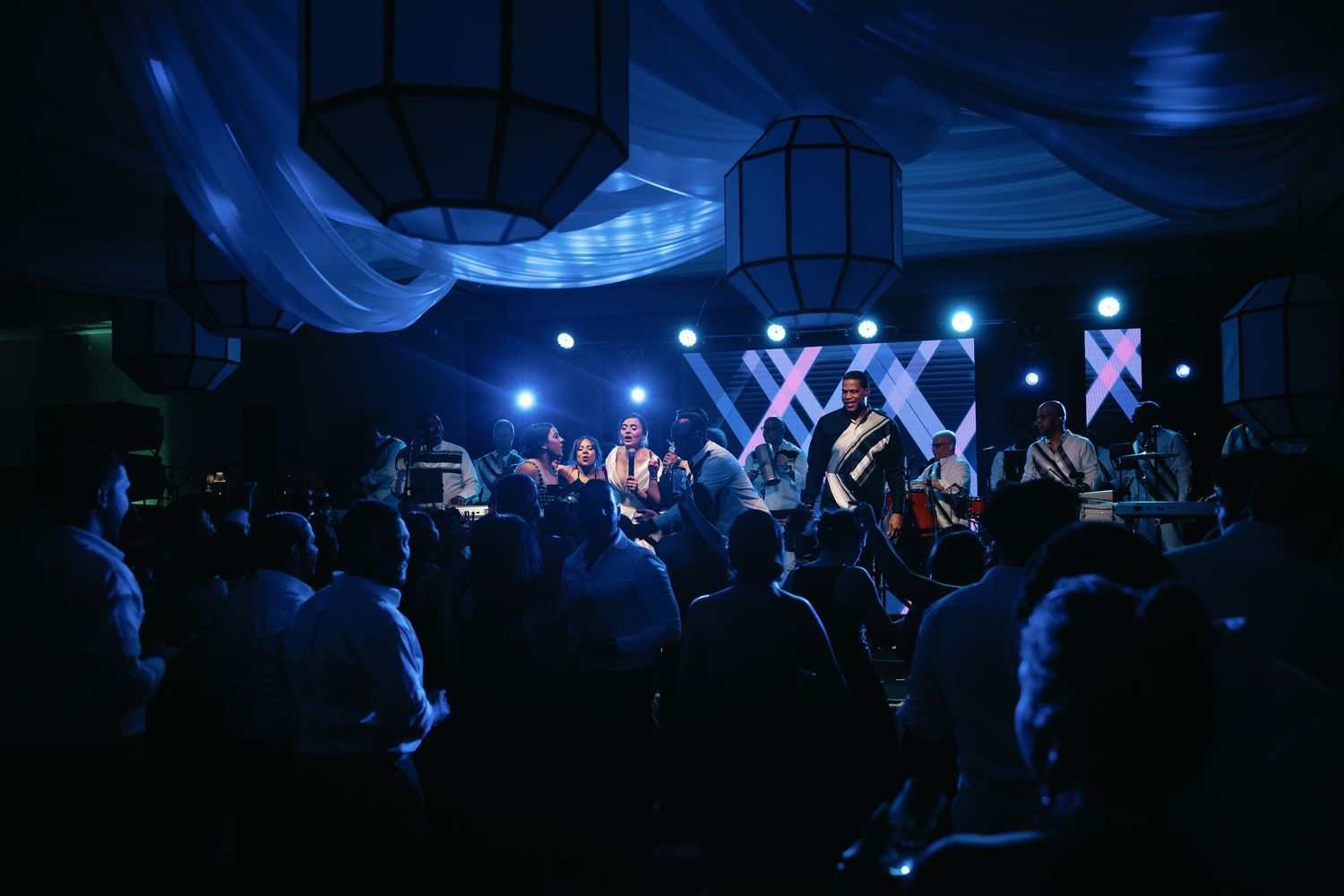Energetic crowd dancing at a nightclub with blue lighting and white fabric decorations overhead.